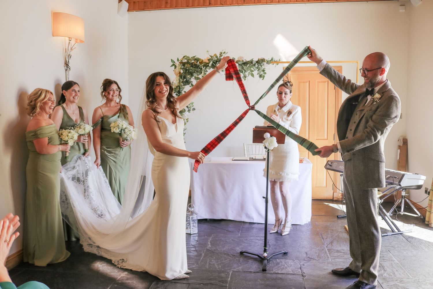 Wedding party stands at altar as officiant performs marriage ceremony with decorative wooden arch and green floral decor.