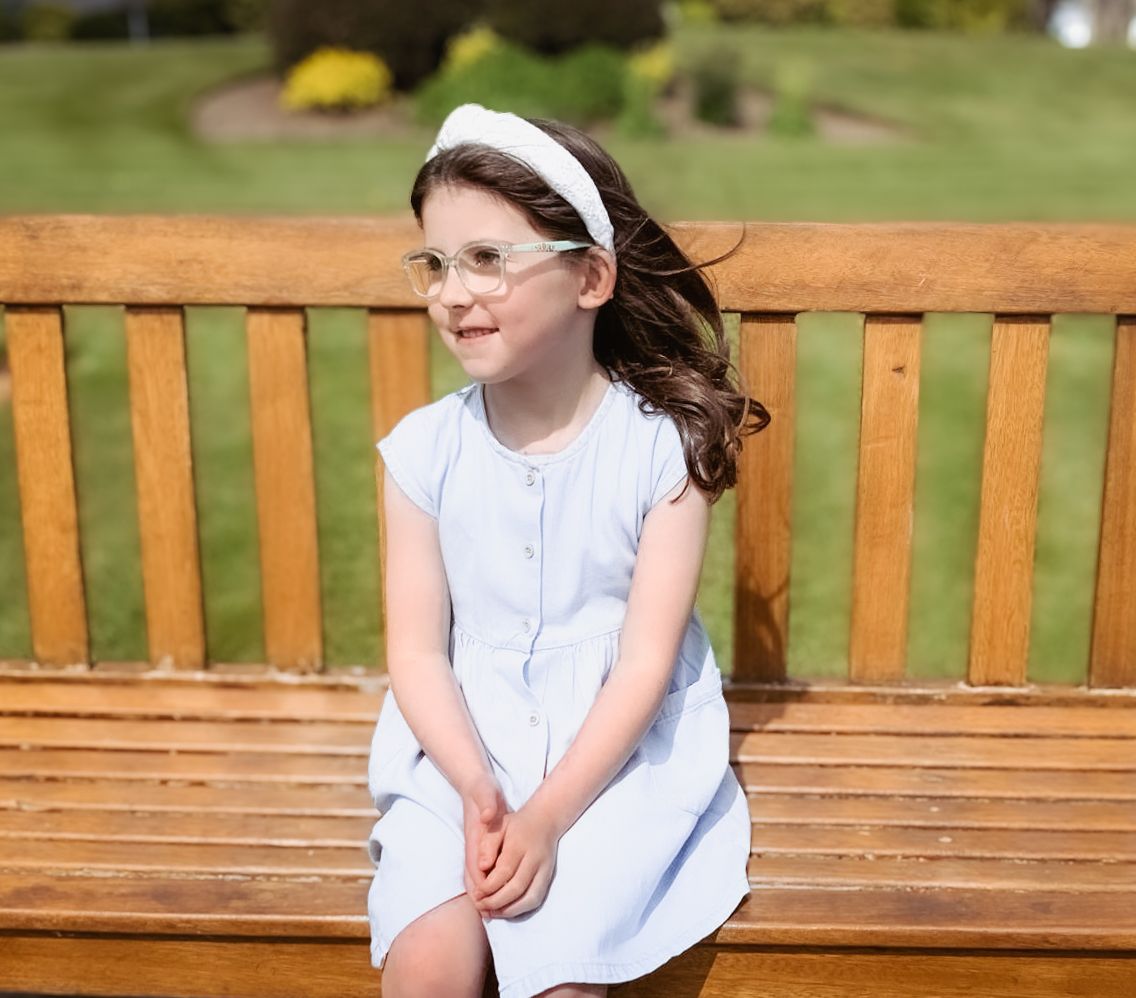 A child in a white dress and headband sits on a wooden park bench while wind blows through their hair.