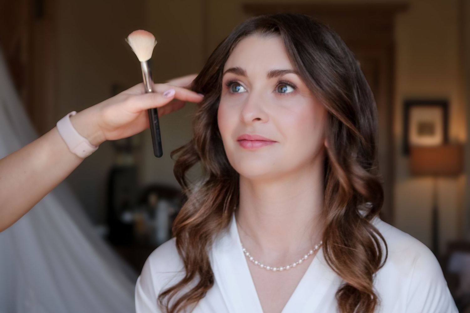A makeup artist applies cosmetic powder with a brush while a person in a white robe and pearl necklace sits patiently.