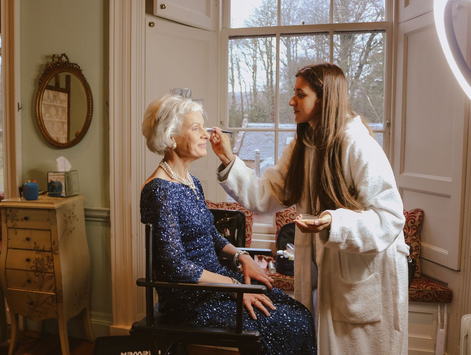 Healthcare worker in white coat provides care to elderly patient sitting in wheelchair by window in home setting.