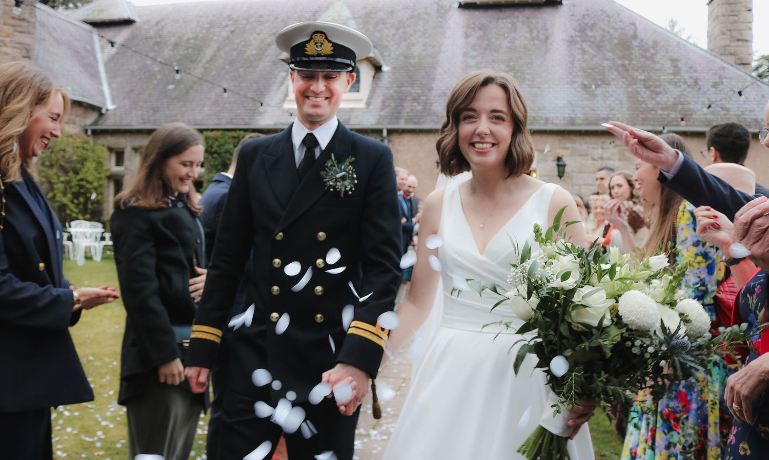 Wedding celebration outdoors with people throwing confetti as uniformed naval officer and bride walk past floral arrangements.