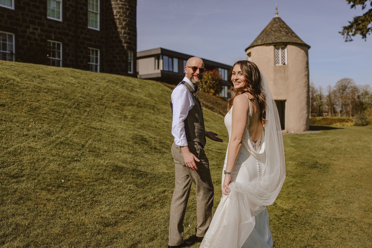 A couple in wedding attire poses on a grassy hillside near a stone tower on their special day.