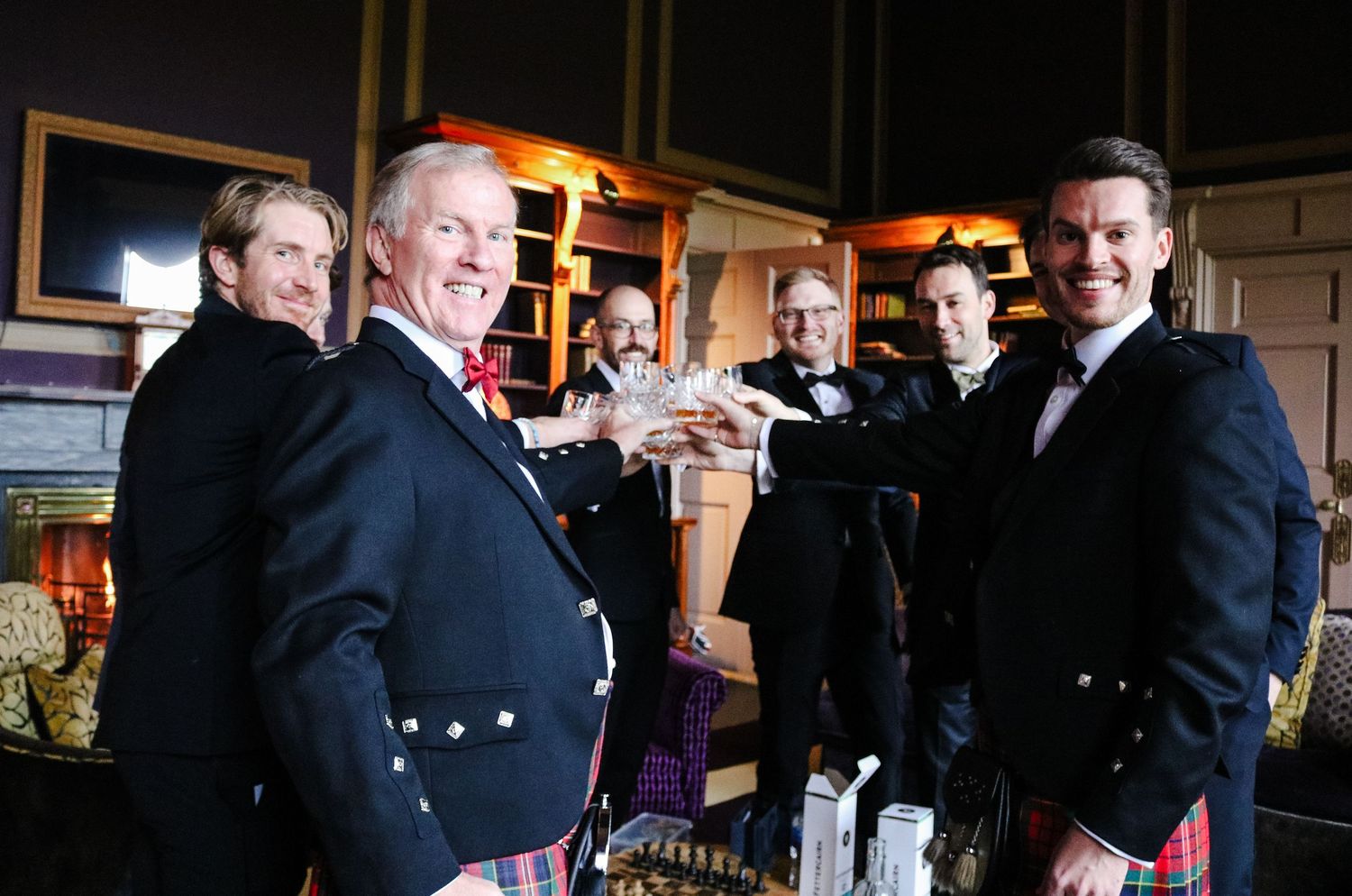 Group of men in traditional Scottish kilts toasting with drinks at an indoor gathering.
