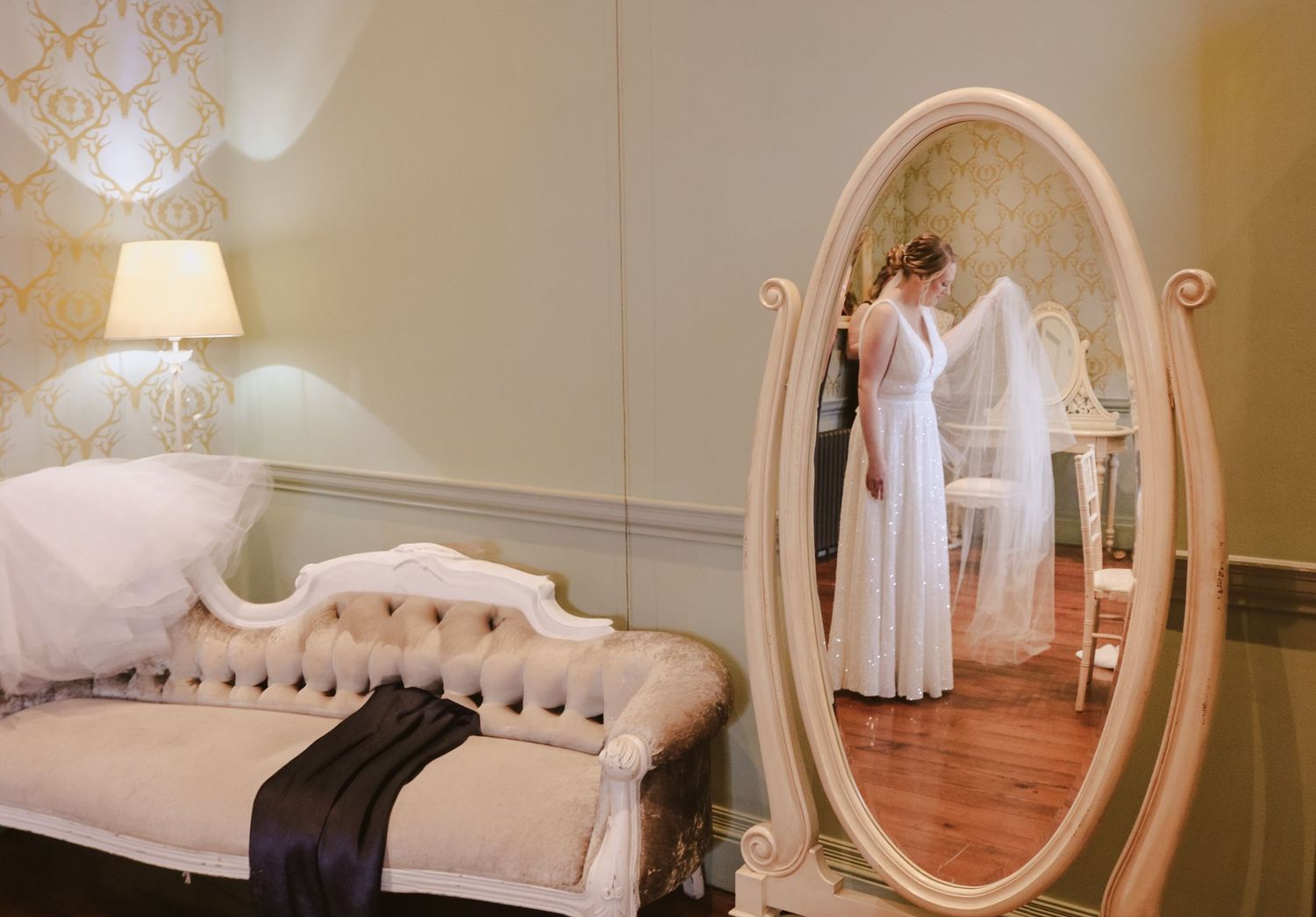 Elegant vintage white dress hangs beside ornate oval mirror in romantic bridal dressing room with plush chaise lounge.