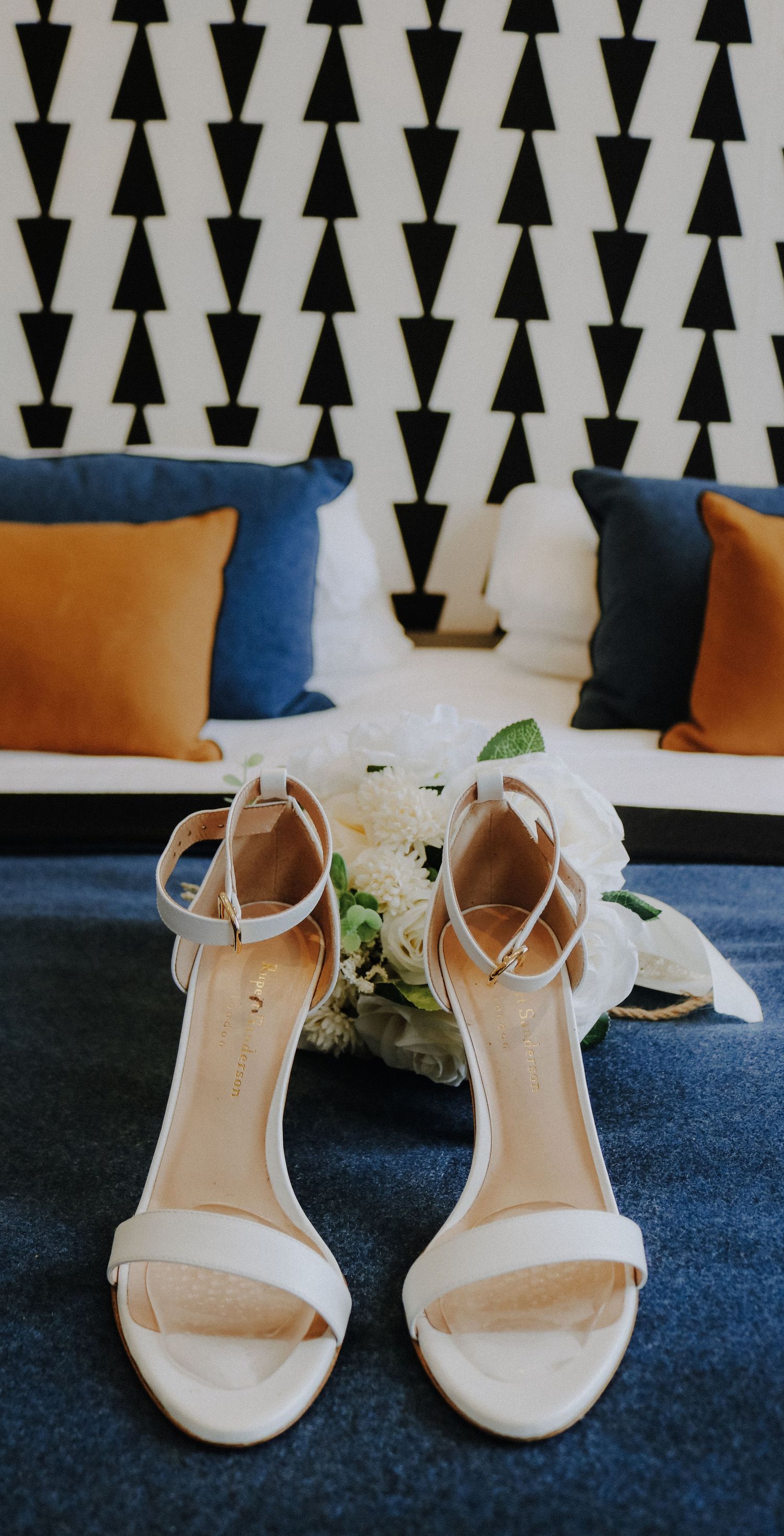 White bridal sandals with ankle straps displayed on blue bedding with geometric pattern backdrop and orange throw pillows.