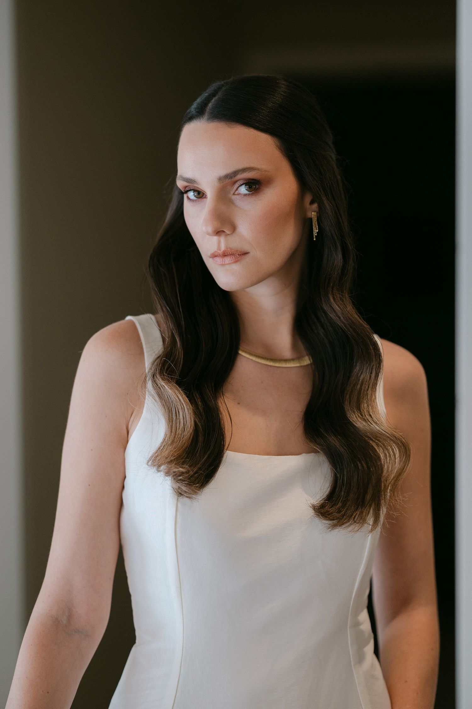 Portrait of a bride in white sleeveless dress with dark wavy hair and minimal makeup in moody lighting.