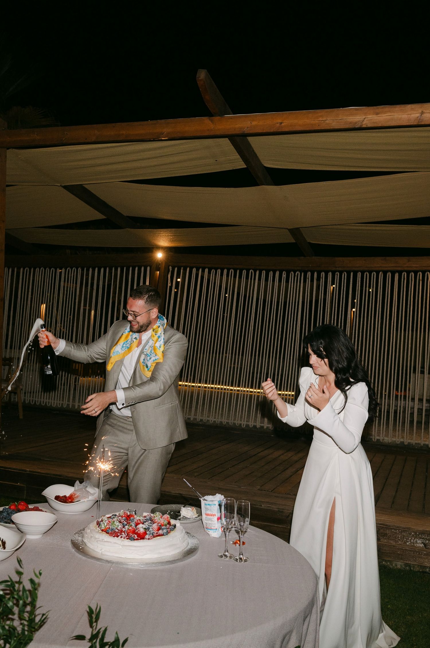 A couple celebrates with sparklers while cutting their wedding cake at an outdoor evening reception.