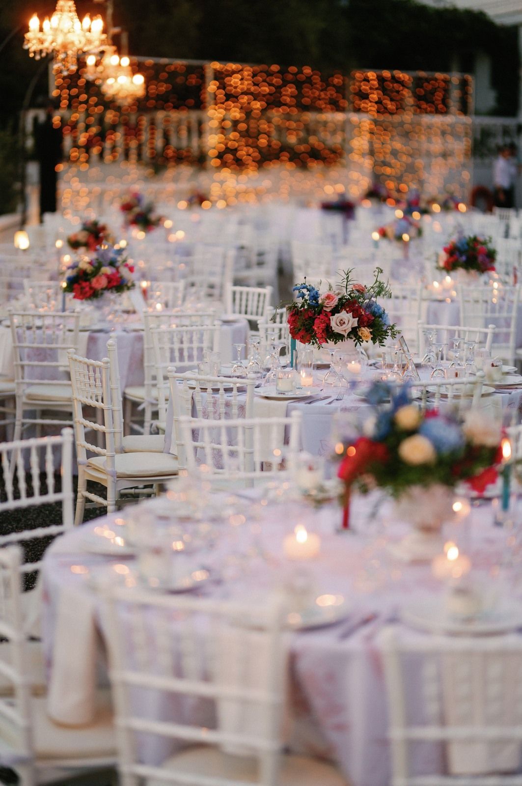 Romantic outdoor wedding reception setup with white tables, chiavari chairs, and floral centerpieces under string lights.
