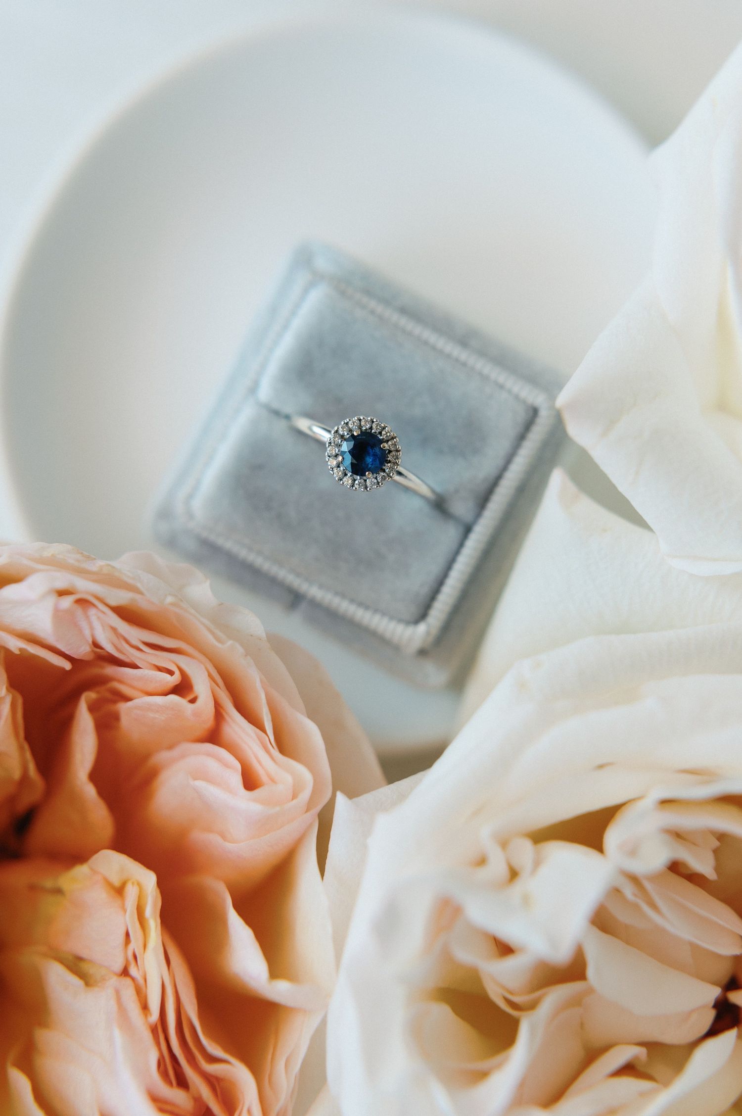 Elegant engagement ring with blue sapphire stone nestled among soft peach and white roses in a romantic setting.