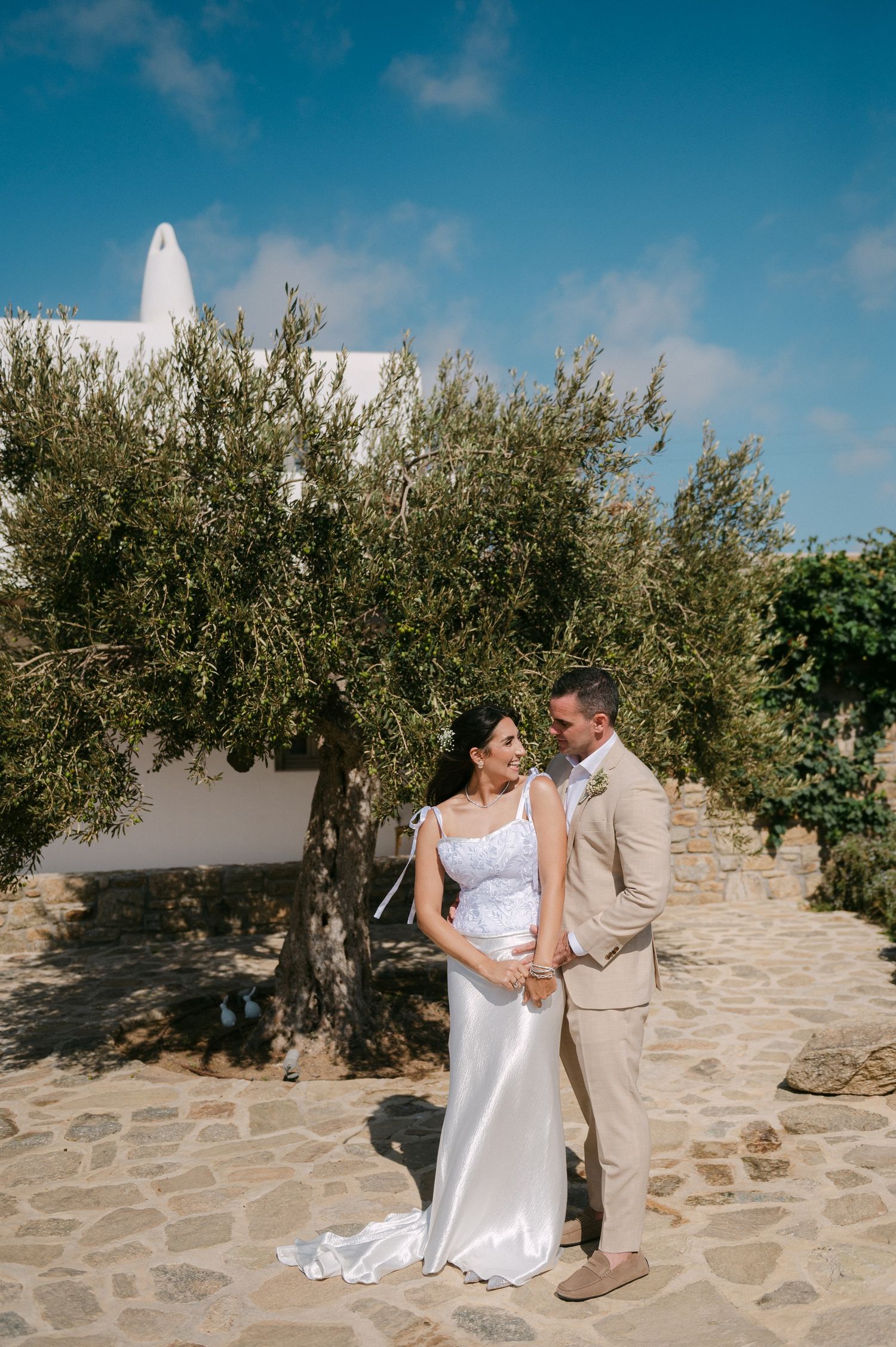 A couple stands together on a stone patio near an olive tree and white Mediterranean building on a sunny day.