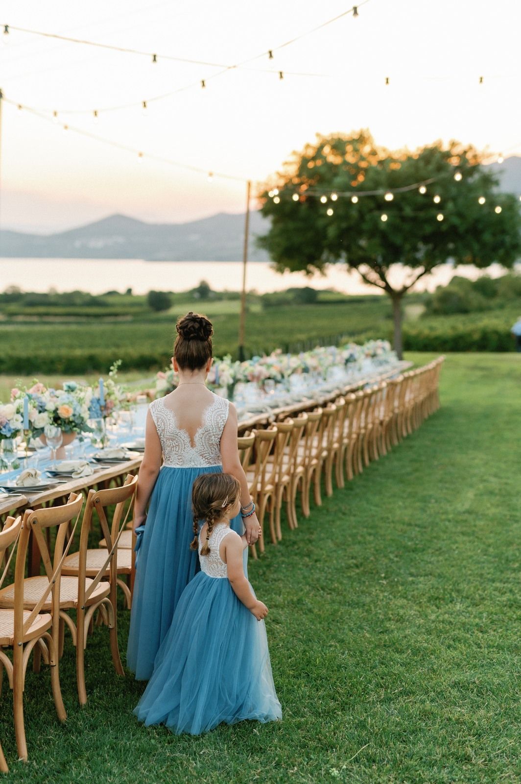 Long outdoor reception table with wooden chairs and string lights set against mountain landscape at sunset.