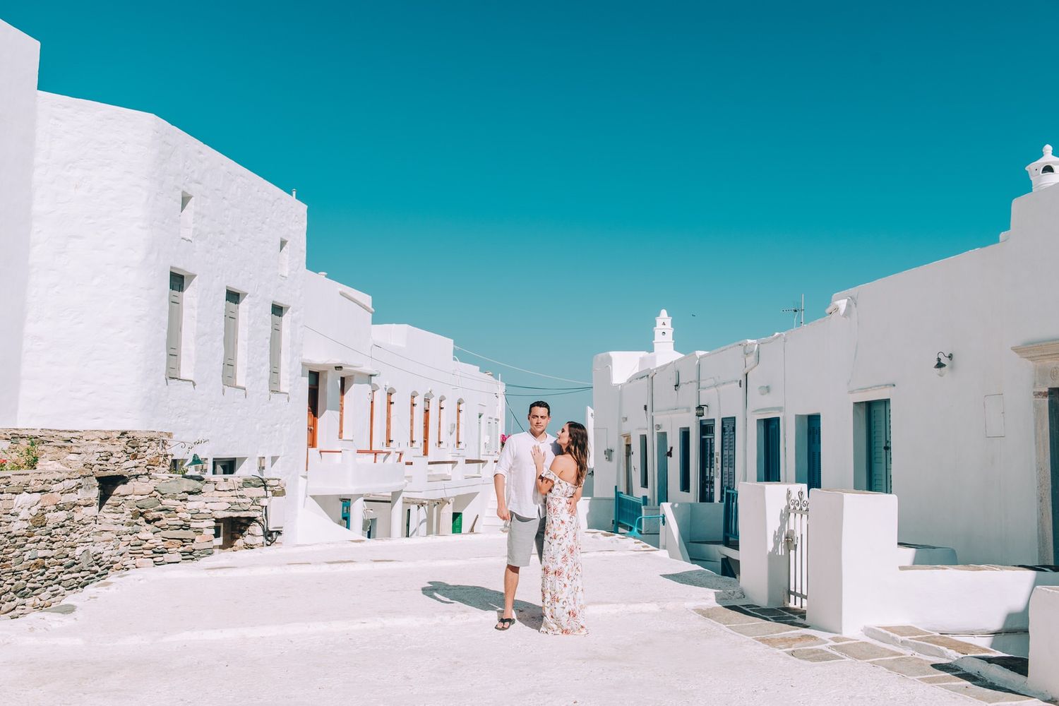 A sun-drenched white street with traditional Cycladic architecture against a vivid blue sky.