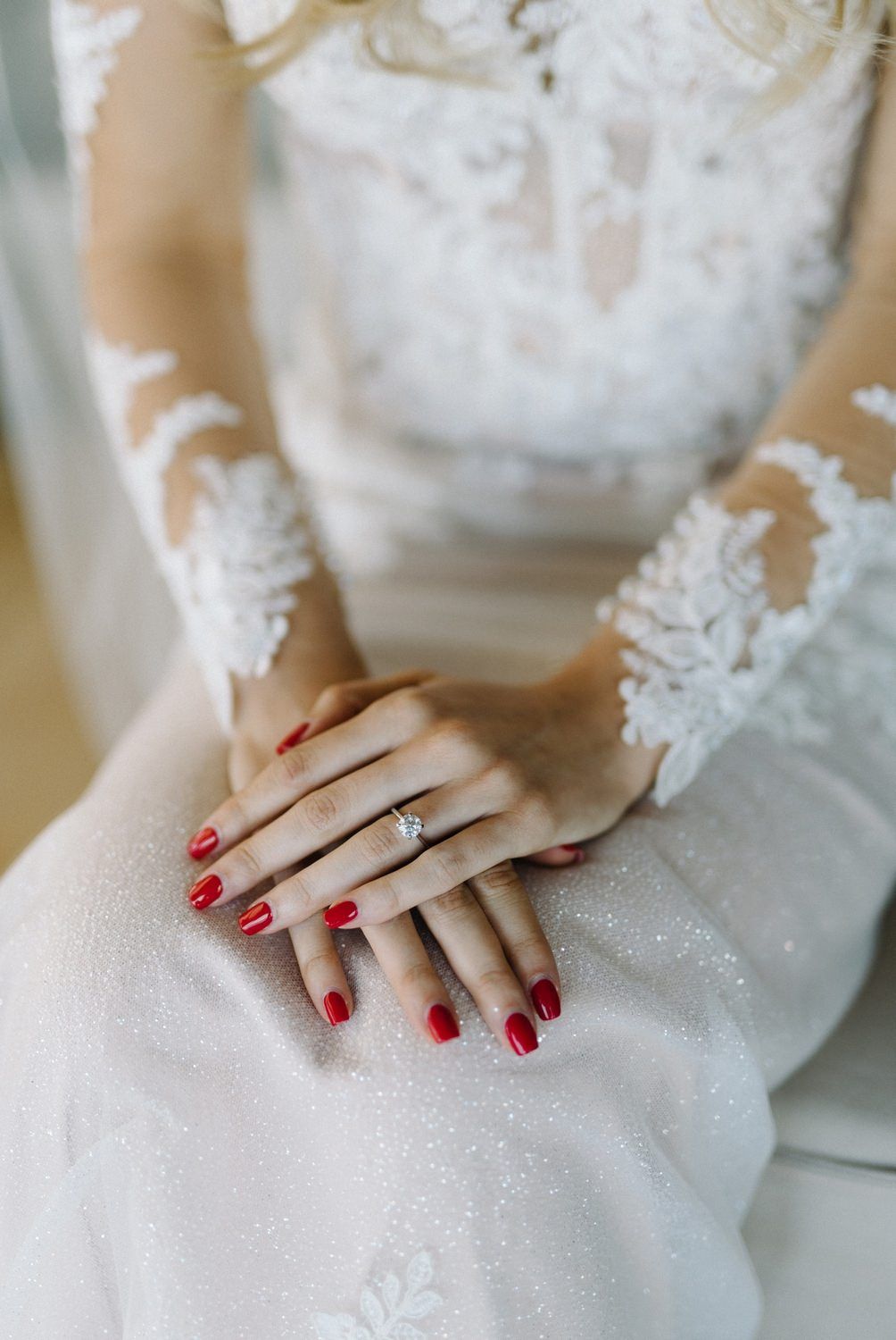 Close-up of hands with red manicured nails and engagement ring on a white lace wedding dress.