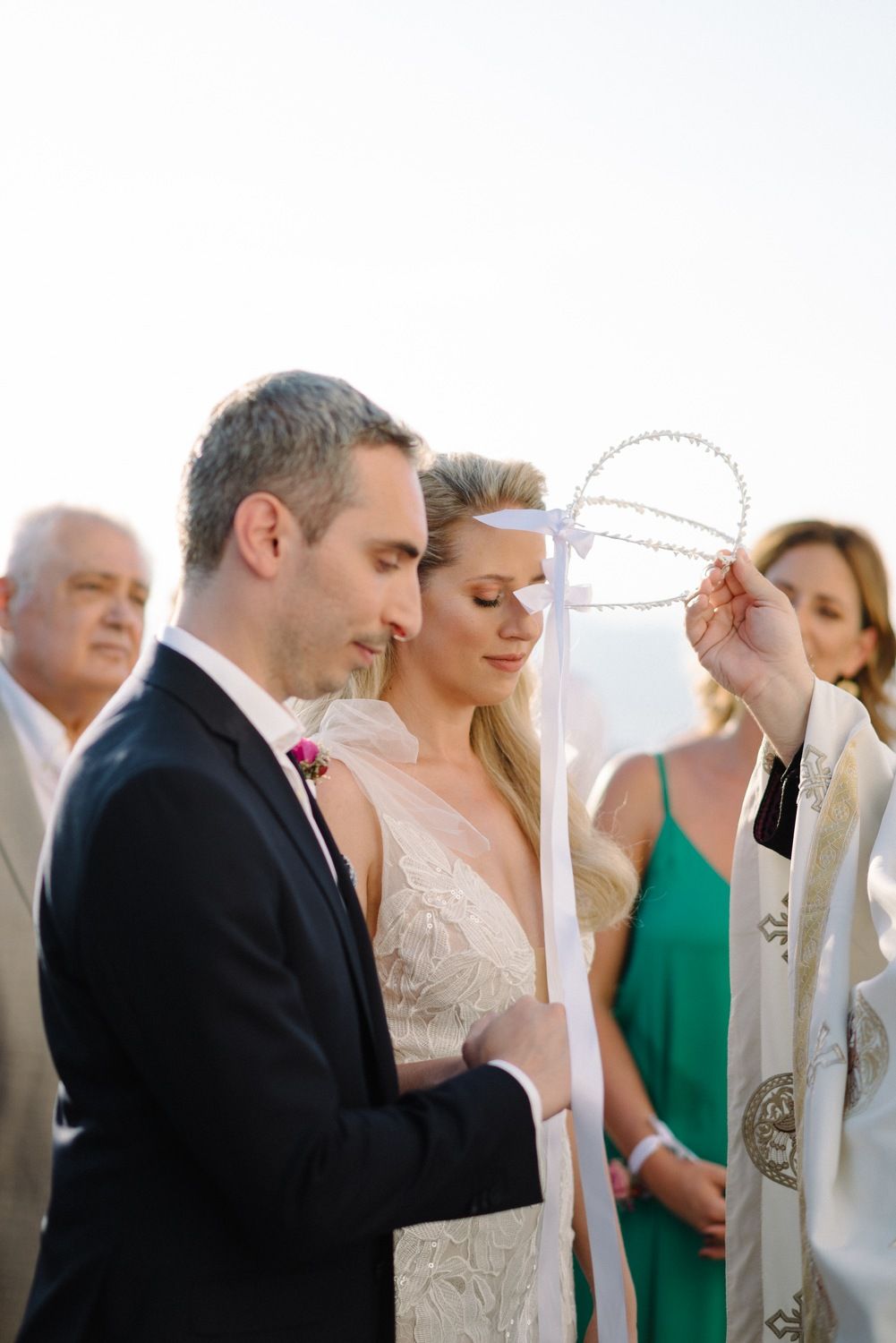 A couple participates in a traditional wedding ceremony with a priest outdoors near the sea.
