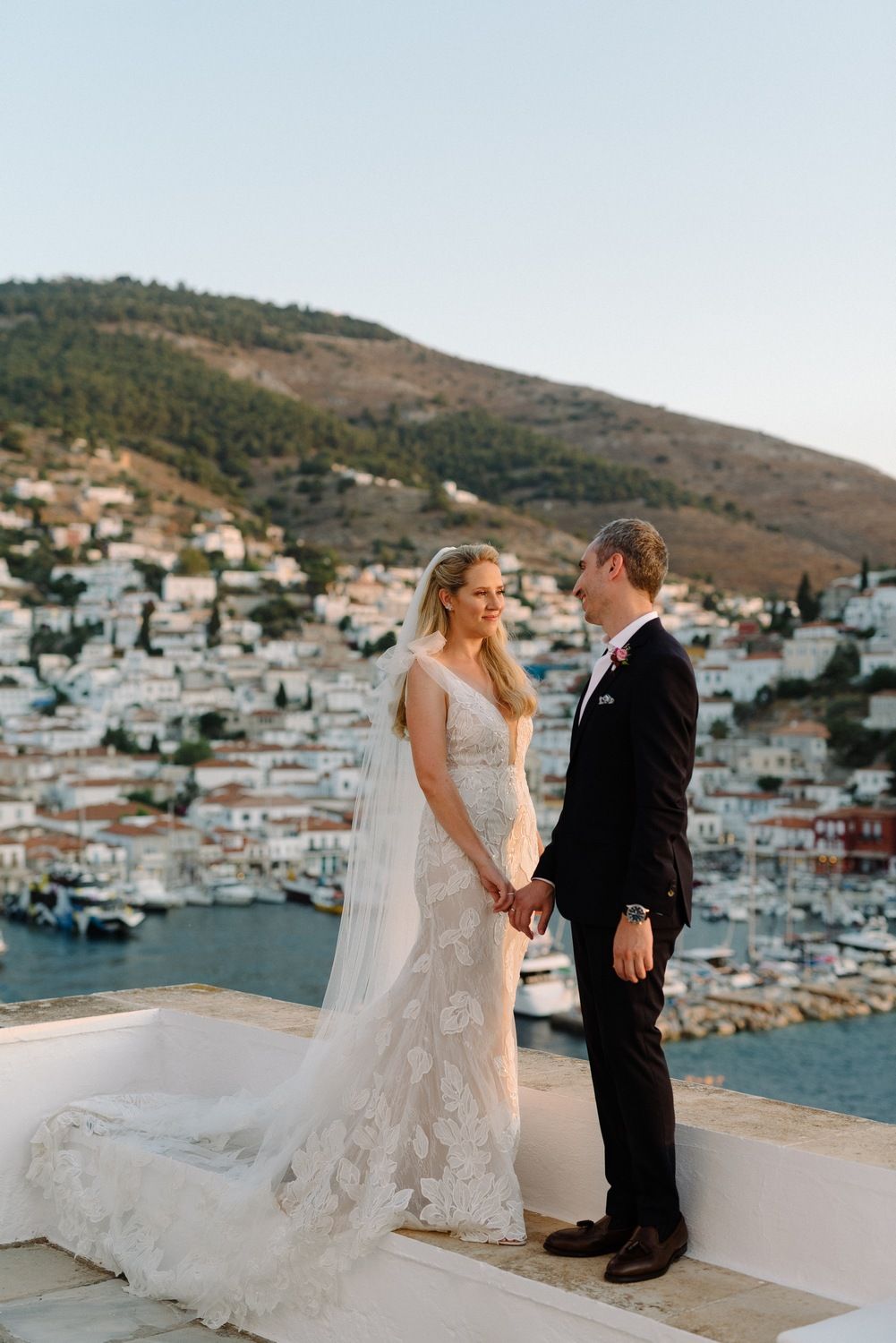 Wedding couple shares intimate moment on balcony overlooking Greek island town and harbor at sunset.