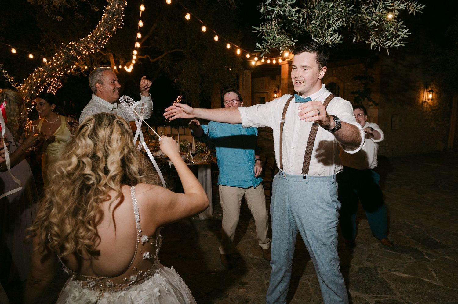 Guests dance joyfully during an evening outdoor wedding reception under twinkling string lights.
