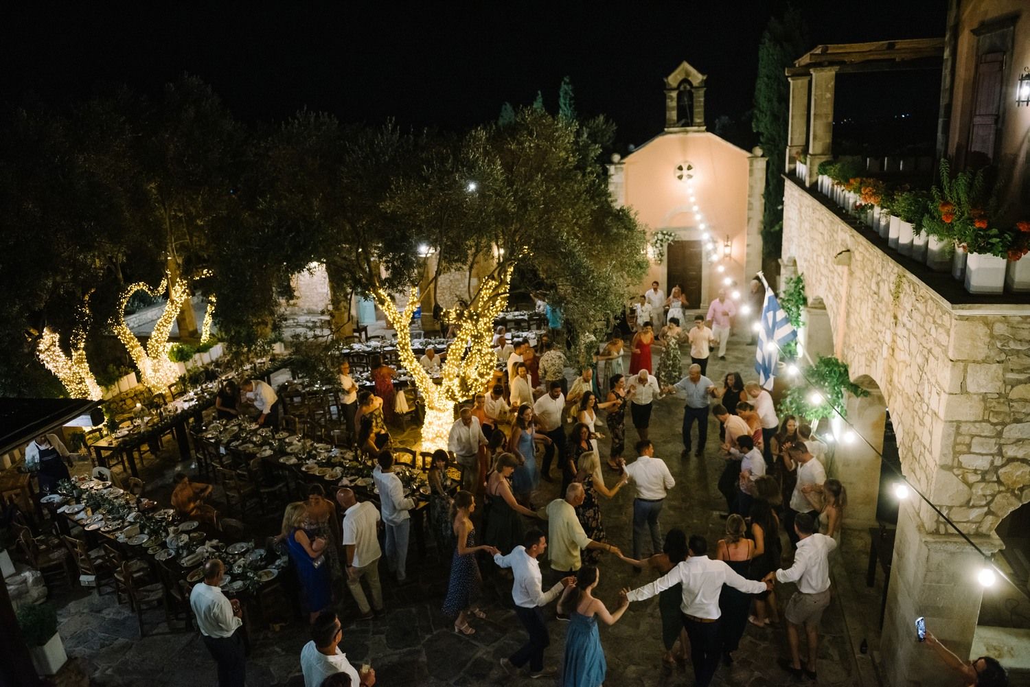 Aerial view of wedding guests dancing in circles under illuminated trees at night.