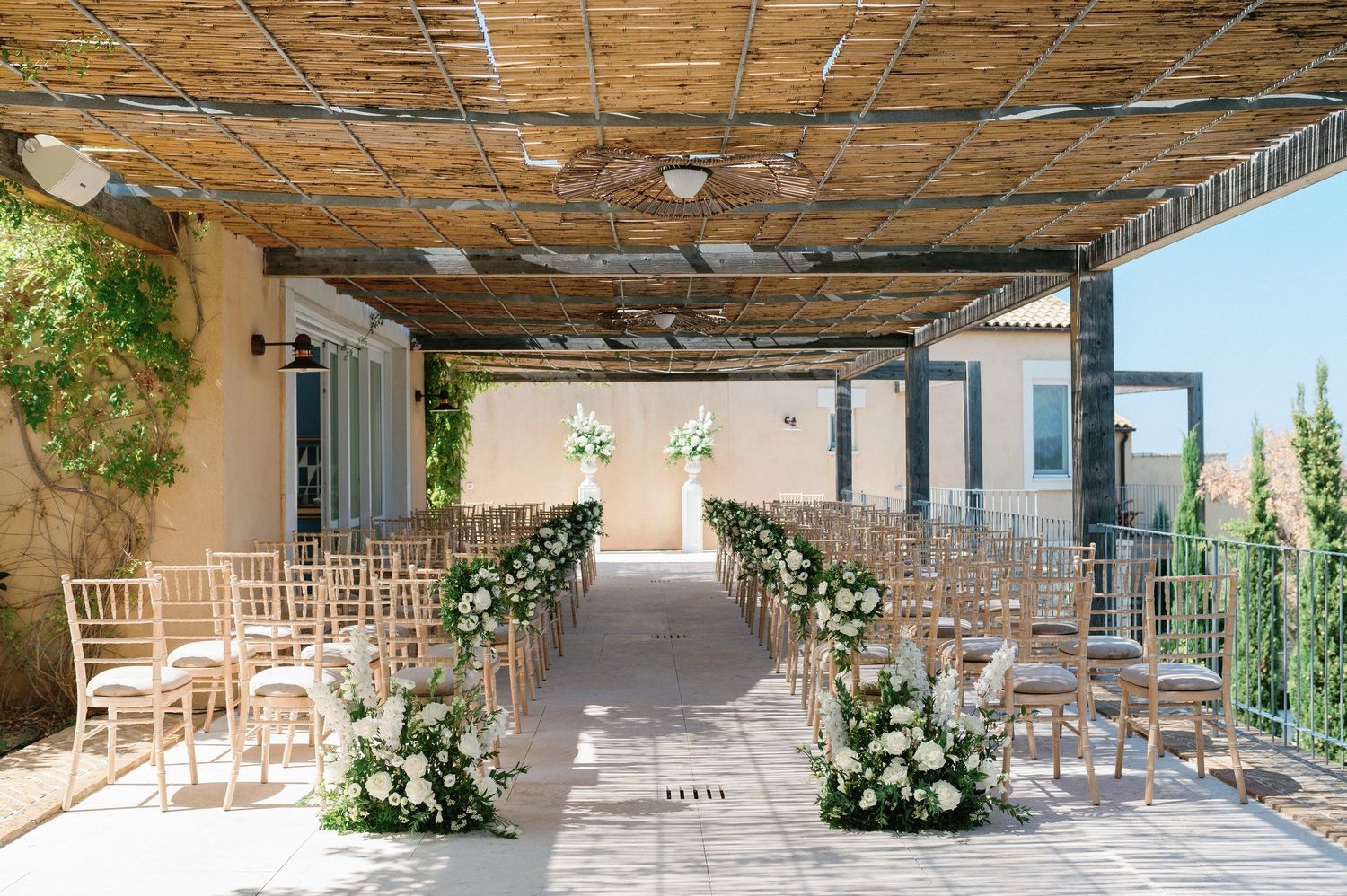An outdoor wedding ceremony setup with white chairs and floral arrangements under a covered walkway.