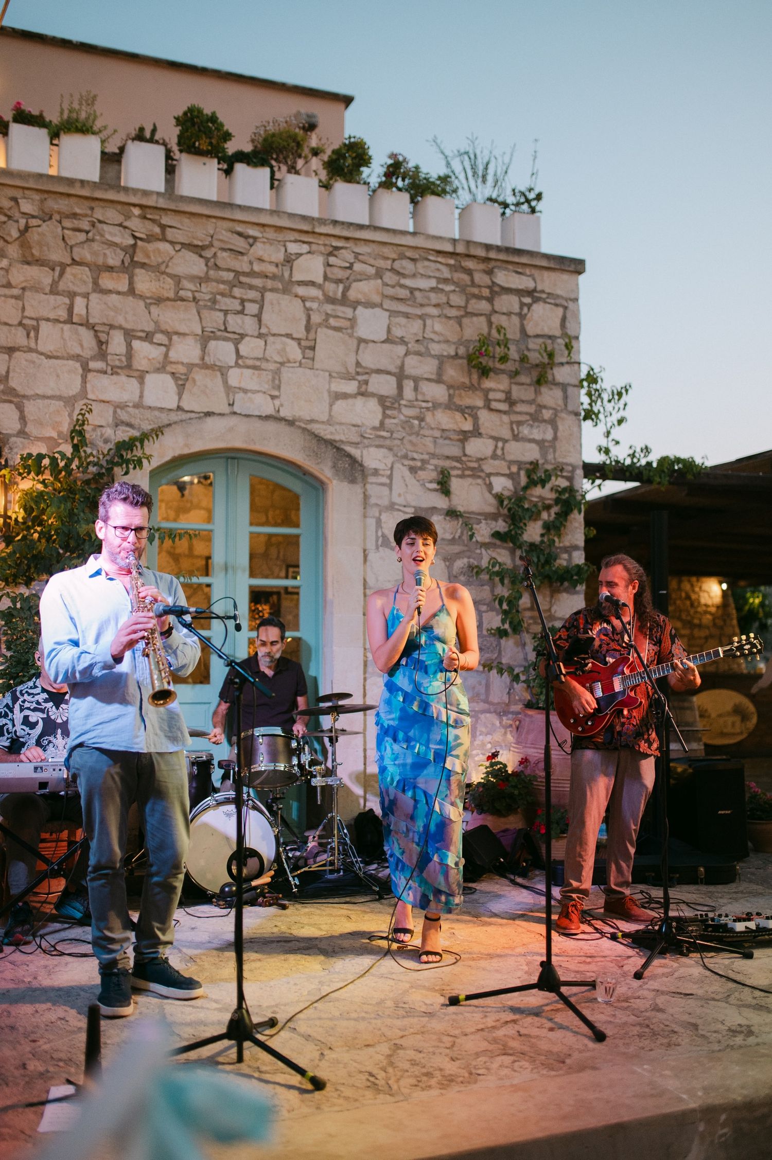 Musicians performing on an outdoor stone patio stage during an evening concert series at a venue.