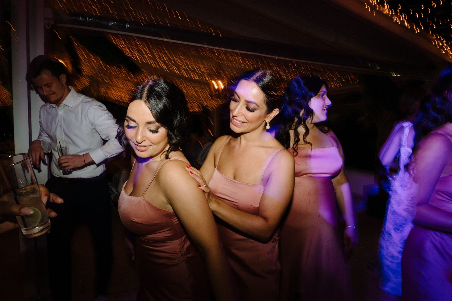 Wedding party members in pink dresses dance together under purple mood lighting.