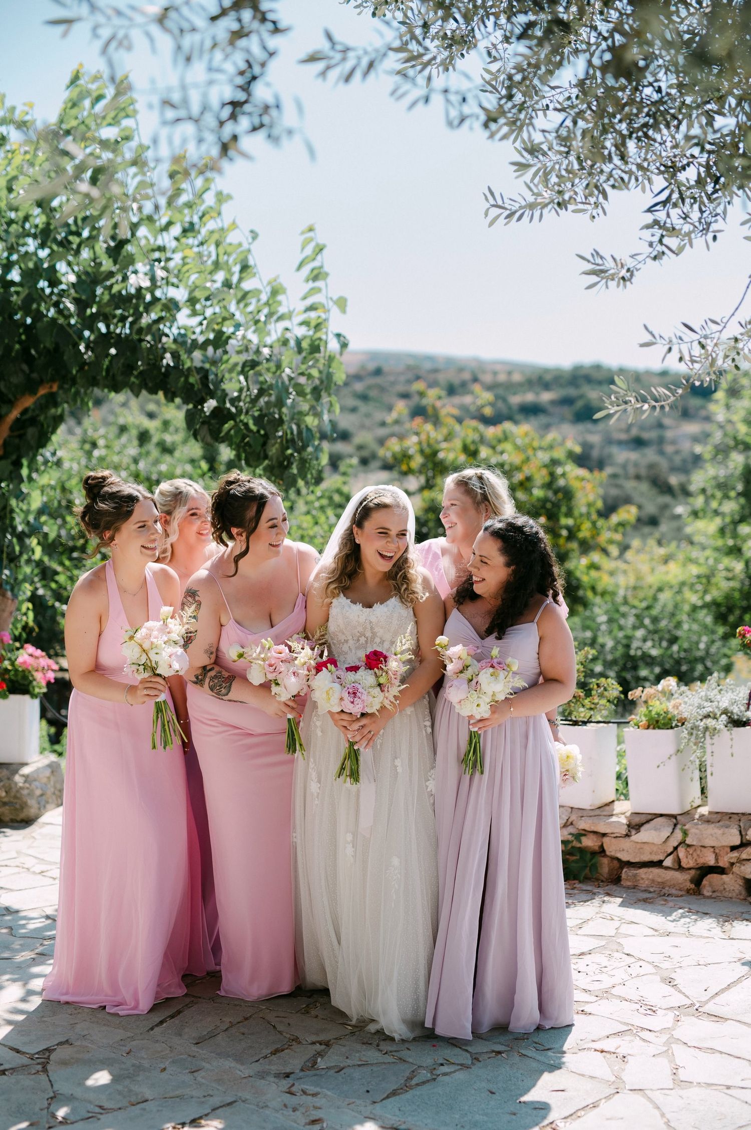Bridesmaids in pink and lavender flowing dresses share a joyful moment on a stone patio with Mediterranean garden backdrop.