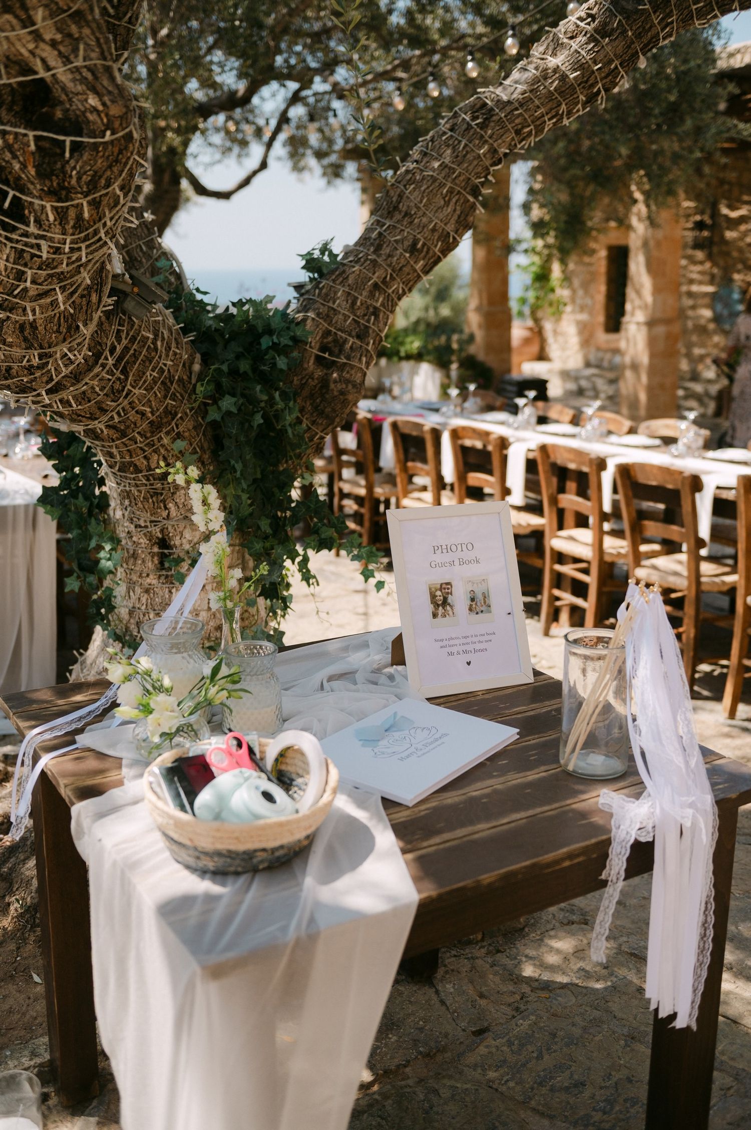 White wedding gift table setup under shady trees with rustic wooden decor and welcome signs at an outdoor venue.