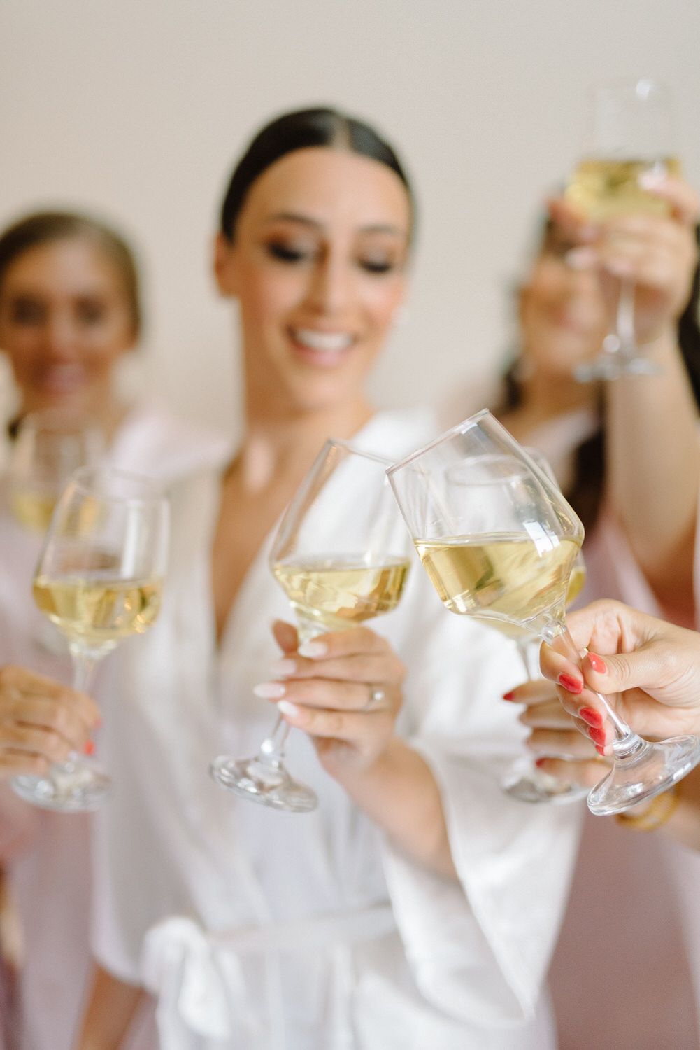 Women in white robes toast with glasses of white wine during pre-wedding celebrations.