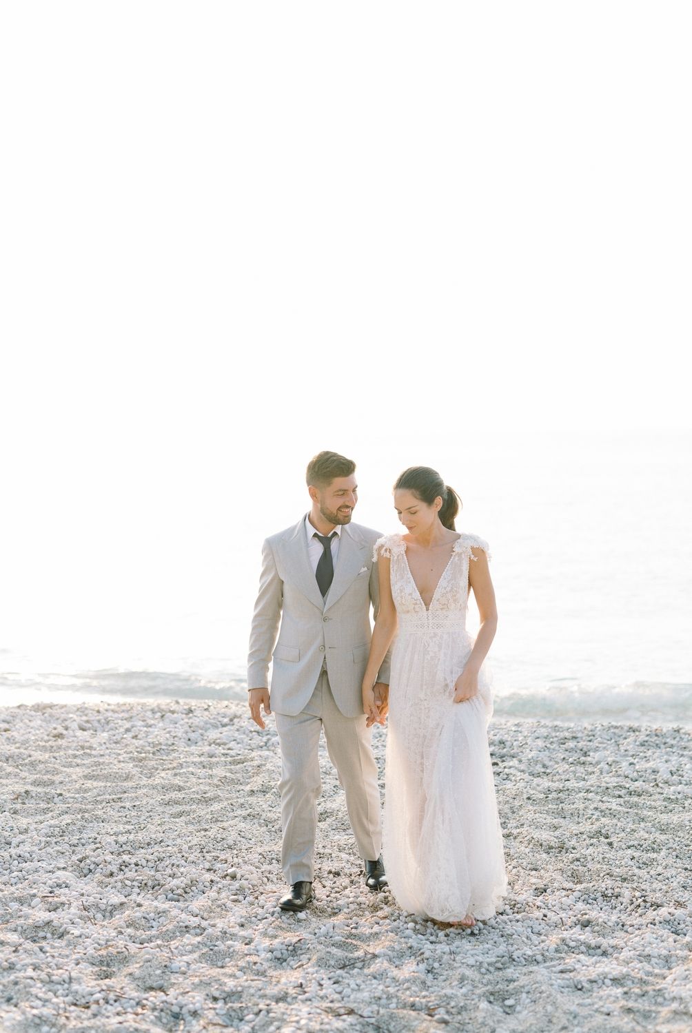 Elegant wedding couple walking together on sandy beach in light-colored formal attire.