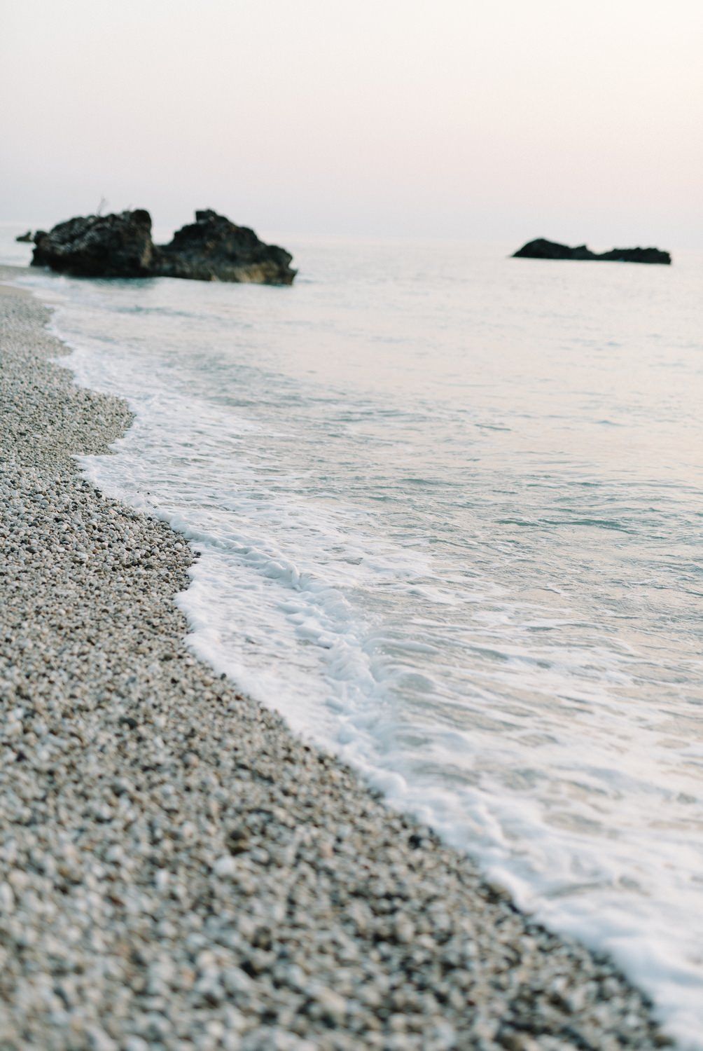 A serene beach landscape with pebbles and dark rocky formations emerging from calm waters.