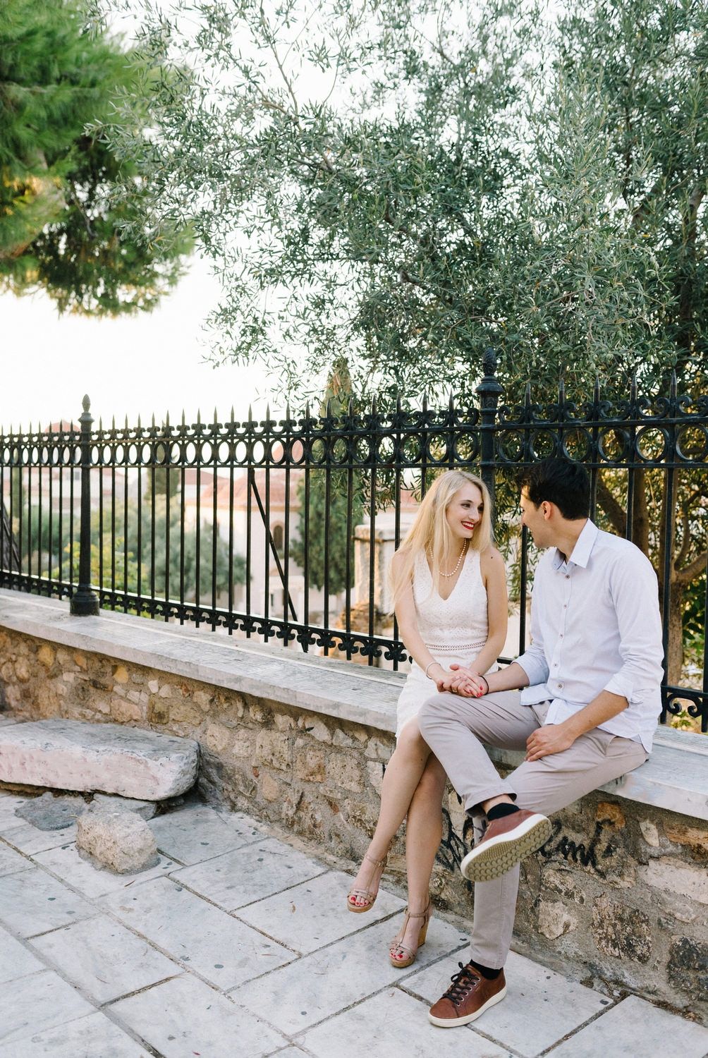 A couple sitting together on a stone wall with Mediterranean scenery in the background.