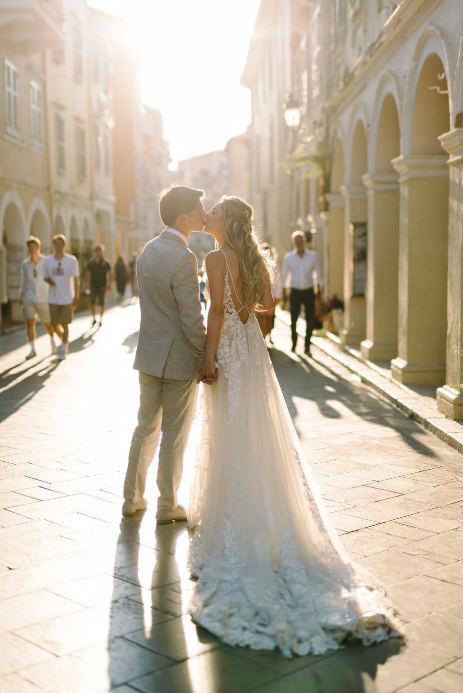 Two newlyweds share an intimate moment on a sunlit street with arched architecture and European charm.