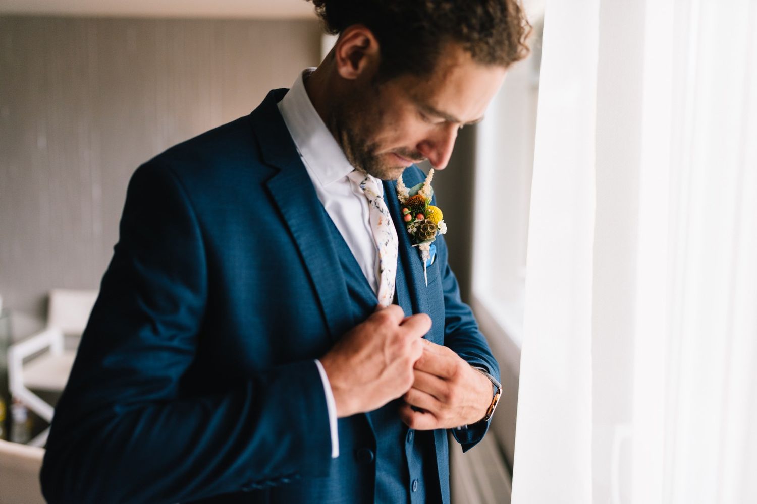 Close-up of groom in navy suit adjusting boutonniere by window light.