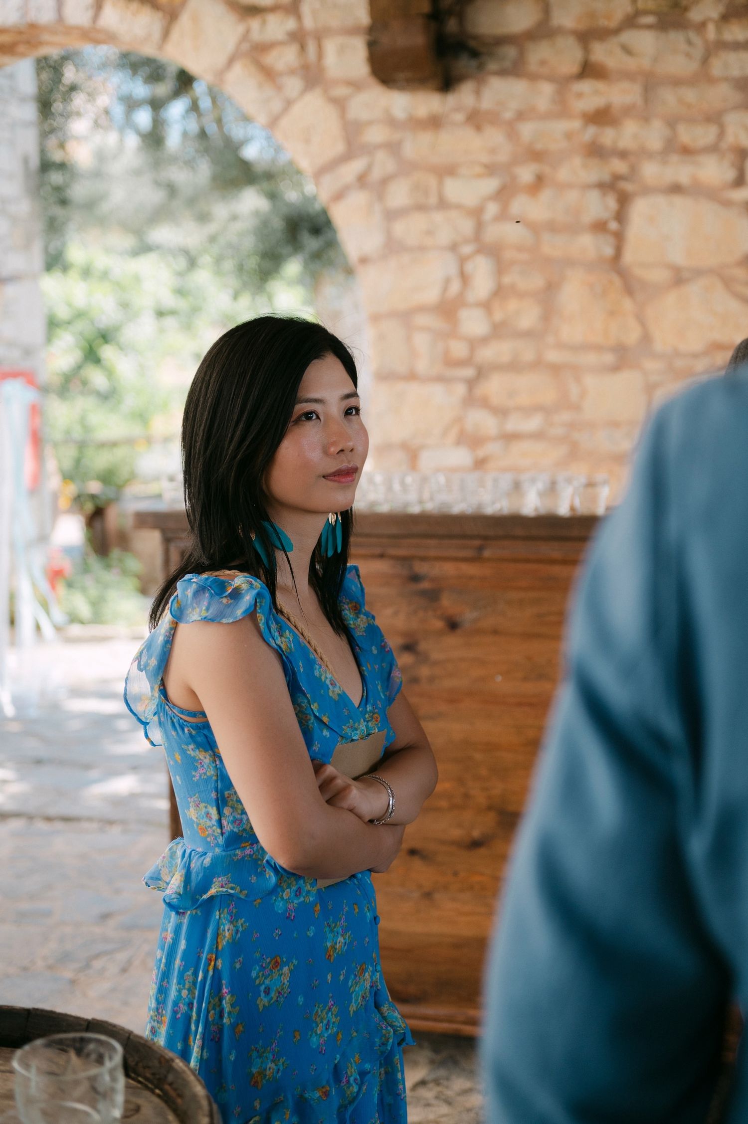 Someone in a blue floral dress stands with arms crossed beneath a stone archway in a rustic setting.