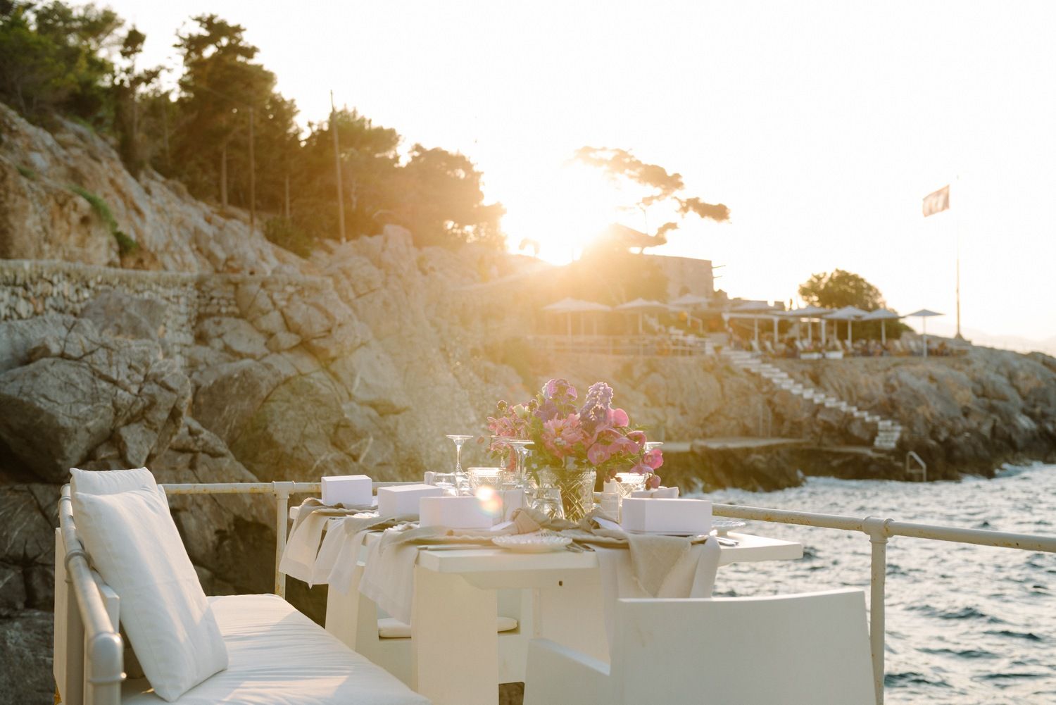 Romantic seaside dinner table setting with floral centerpiece against rocky cliffs at sunset.
