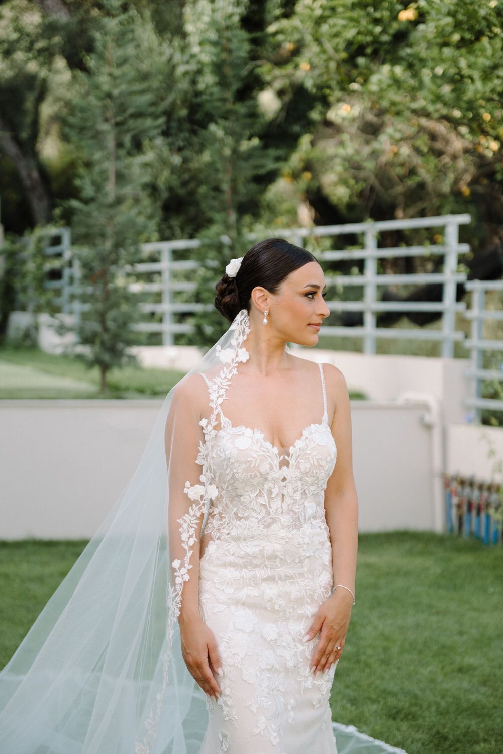 Elegant bride in form-fitting lace wedding gown with long veil poses in garden setting.