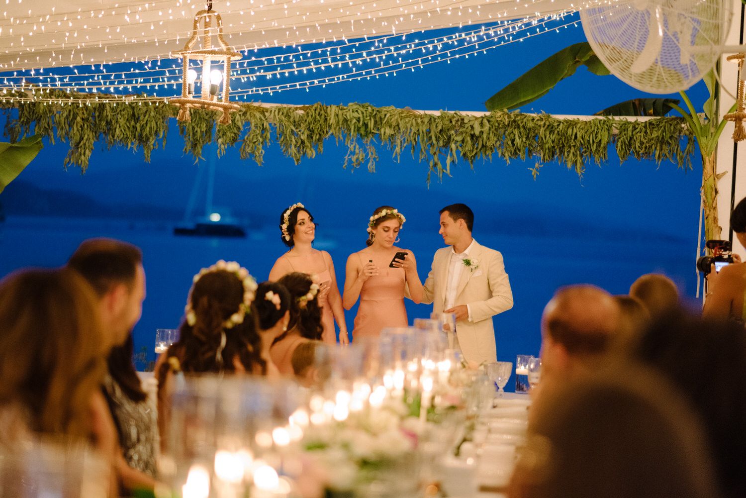 Wedding reception speakers addressing guests at candlelit tables under twinkling lights.