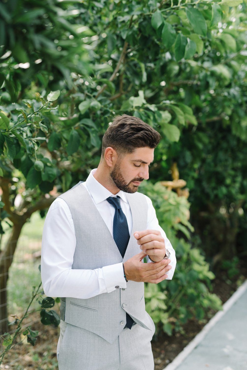 Groom in light gray vest and navy tie adjusting cuff in garden setting.