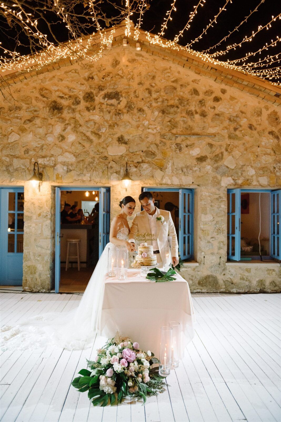 Elegant wedding reception setup with string lights and rustic stone walls featuring white table decor and floral arrangements.