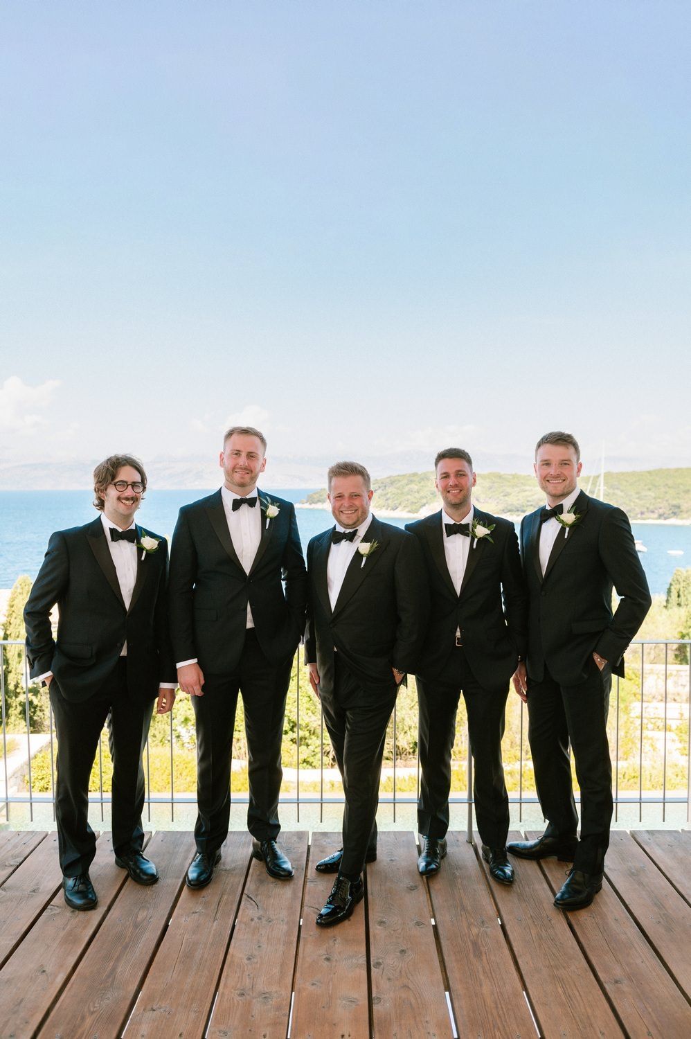 Groomsmen in black tuxedos pose playfully while sitting on a wooden deck at corfu island