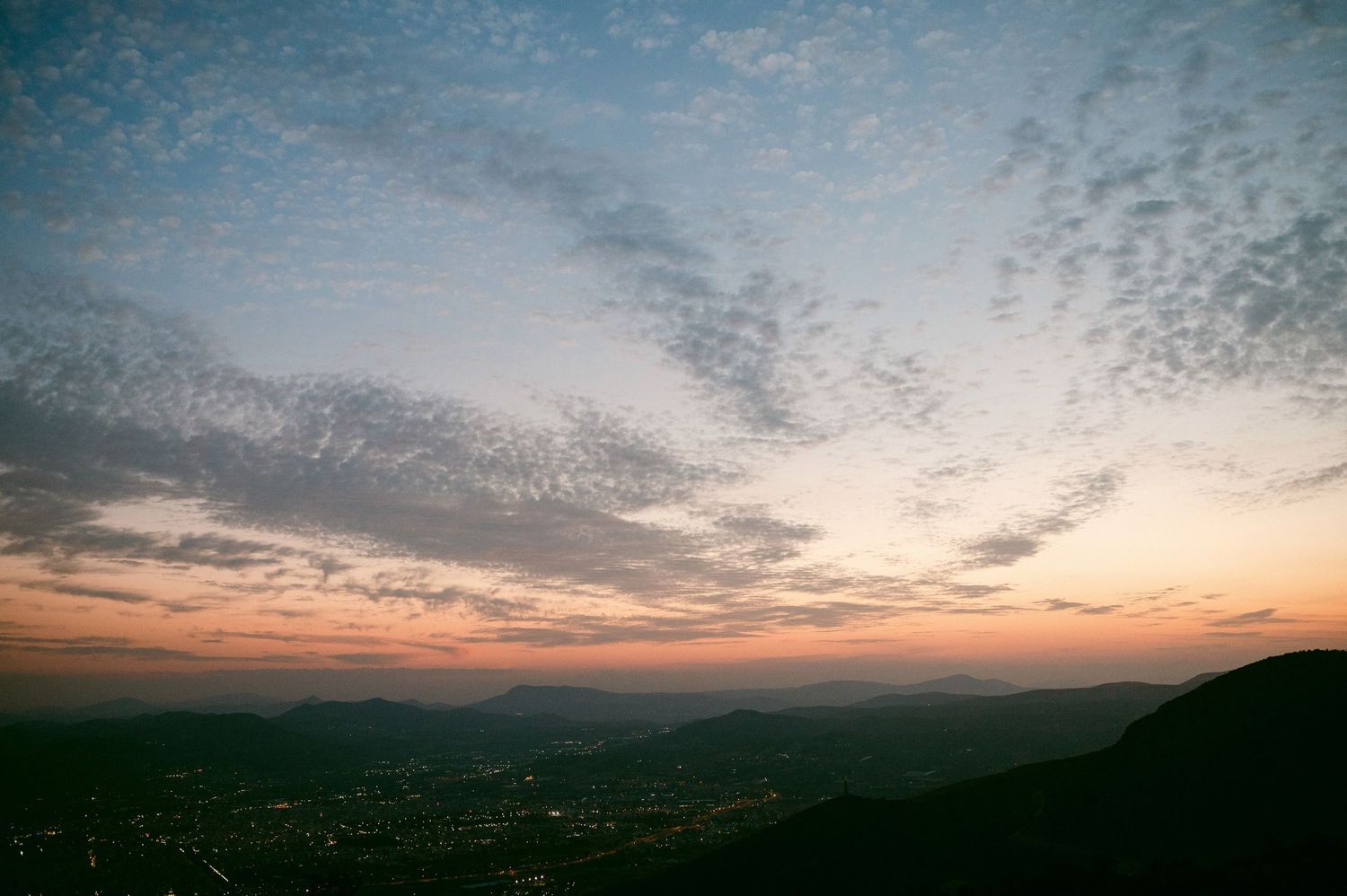 Dramatic sunset over a dark cityscape viewed from hilltop with scattered clouds in orange and blue sky.