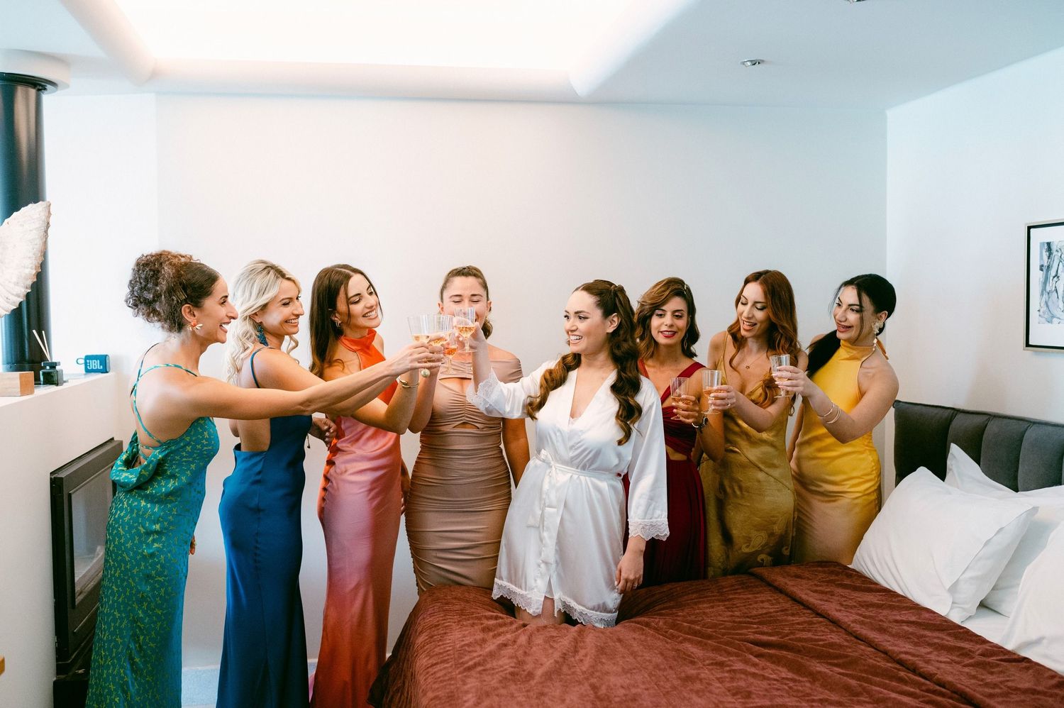 Bridal party in colorful dresses toasting with drinks while celebrating in a hotel room together.