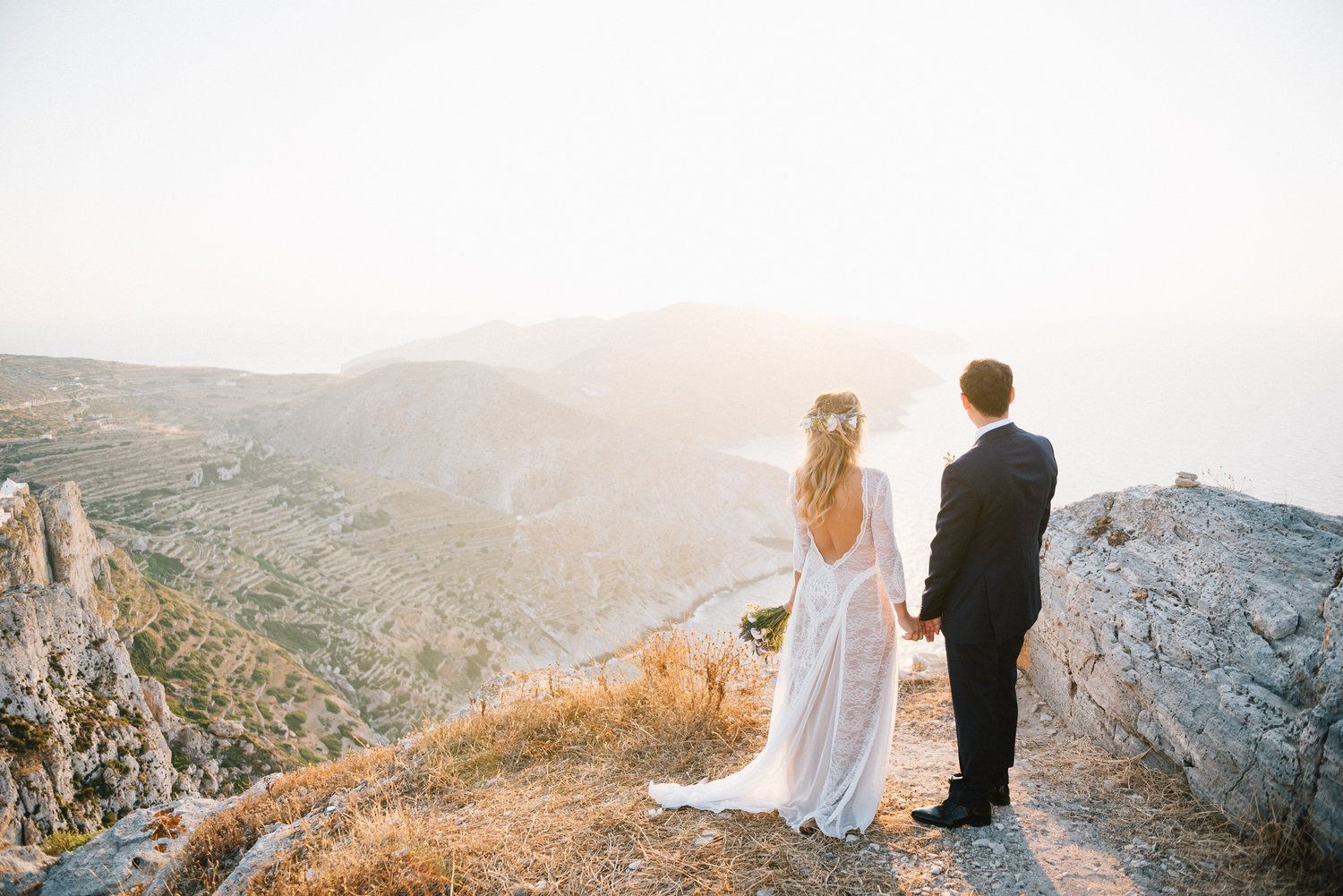 Couple in wedding attire stands on mountain cliff edge at Folegandros island overlooking scenic valley during golden hour sunset.