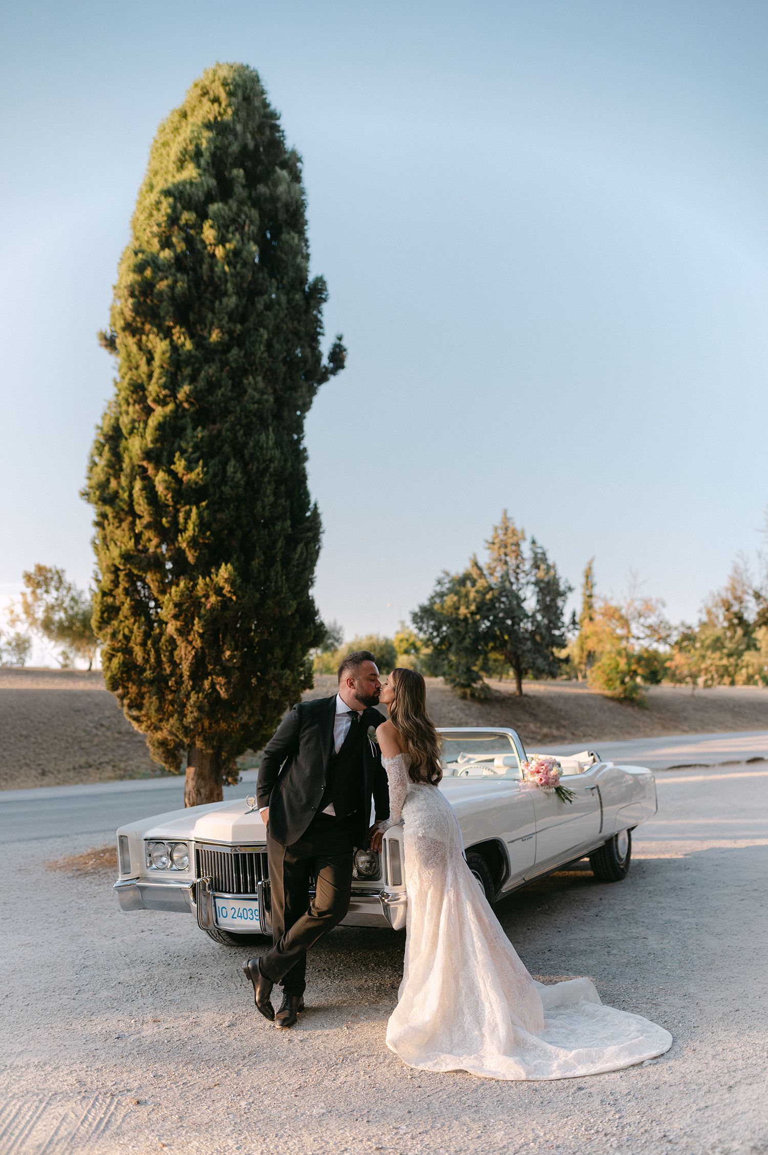 Bride and groom kissing beside a vintage white convertible car with a tall cypress tree backdrop on their wedding day.