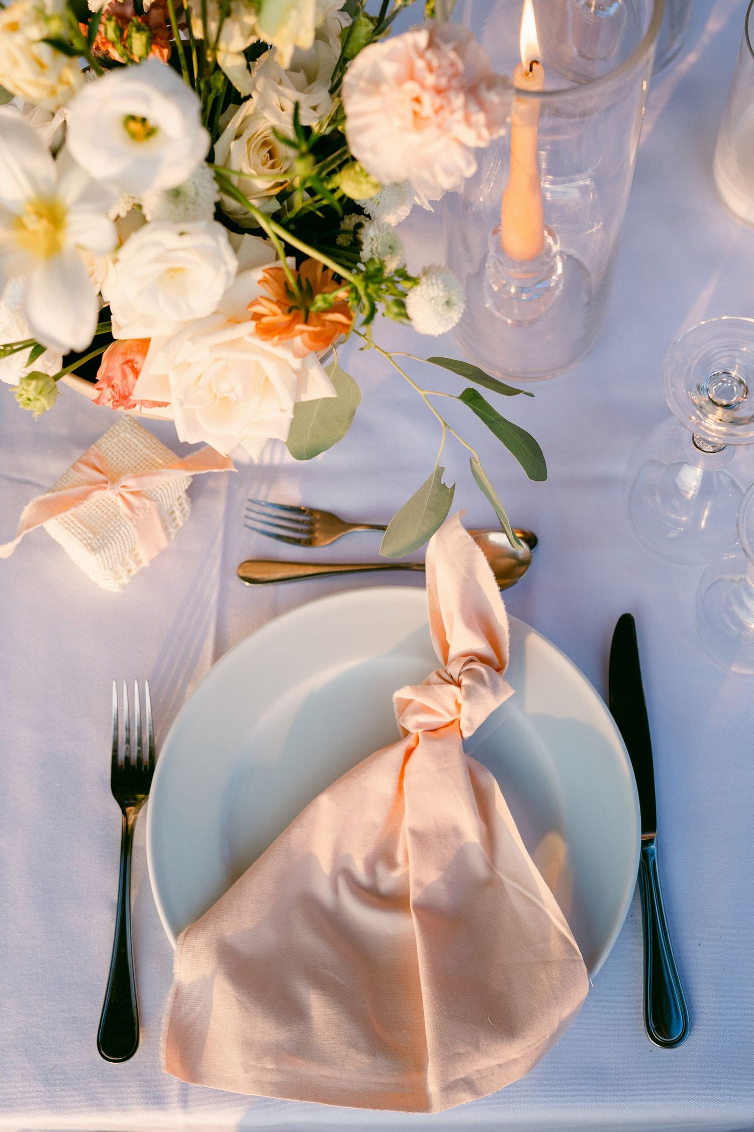 Elegant table setting with peach napkin, white plates, and soft floral centerpiece featuring roses and peonies.