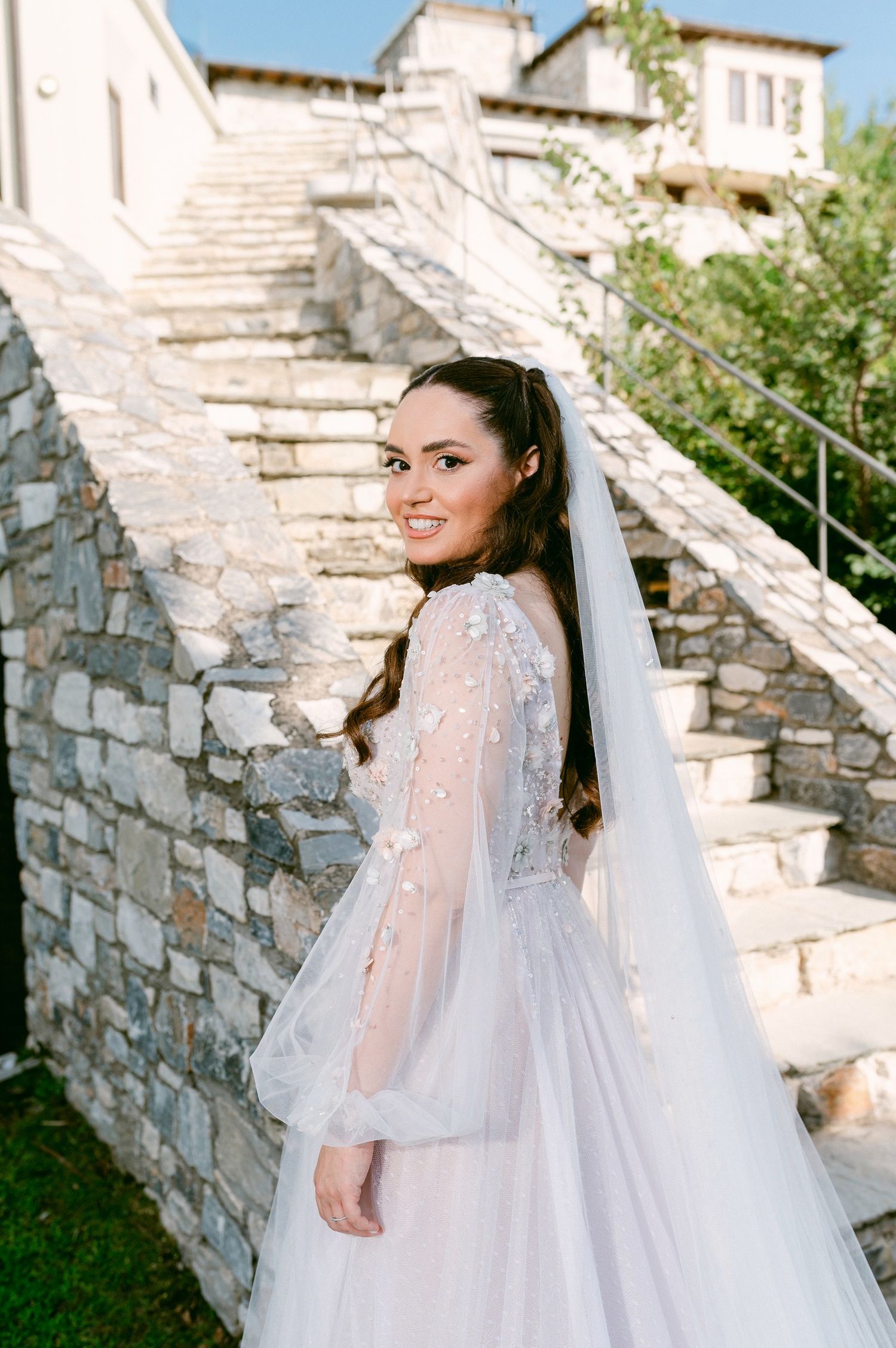 A bride in a flowing long-sleeve lace wedding gown with veil poses on stone steps at an outdoor venue.