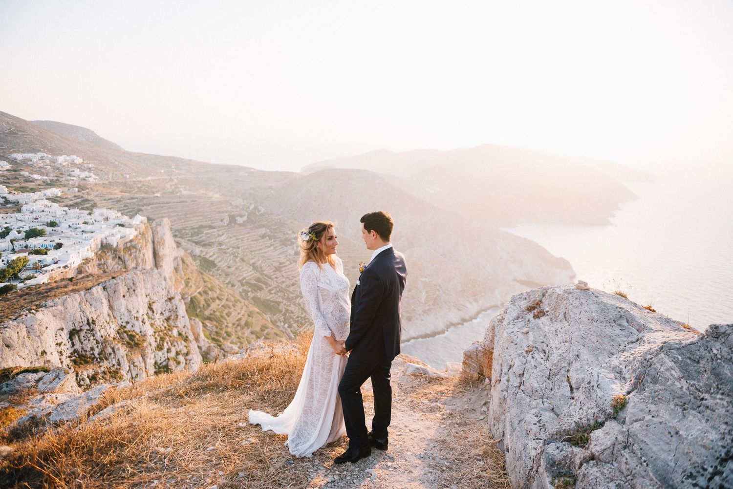 Two people embrace on a mountain cliff during golden hour with scenic coastal views in the background.