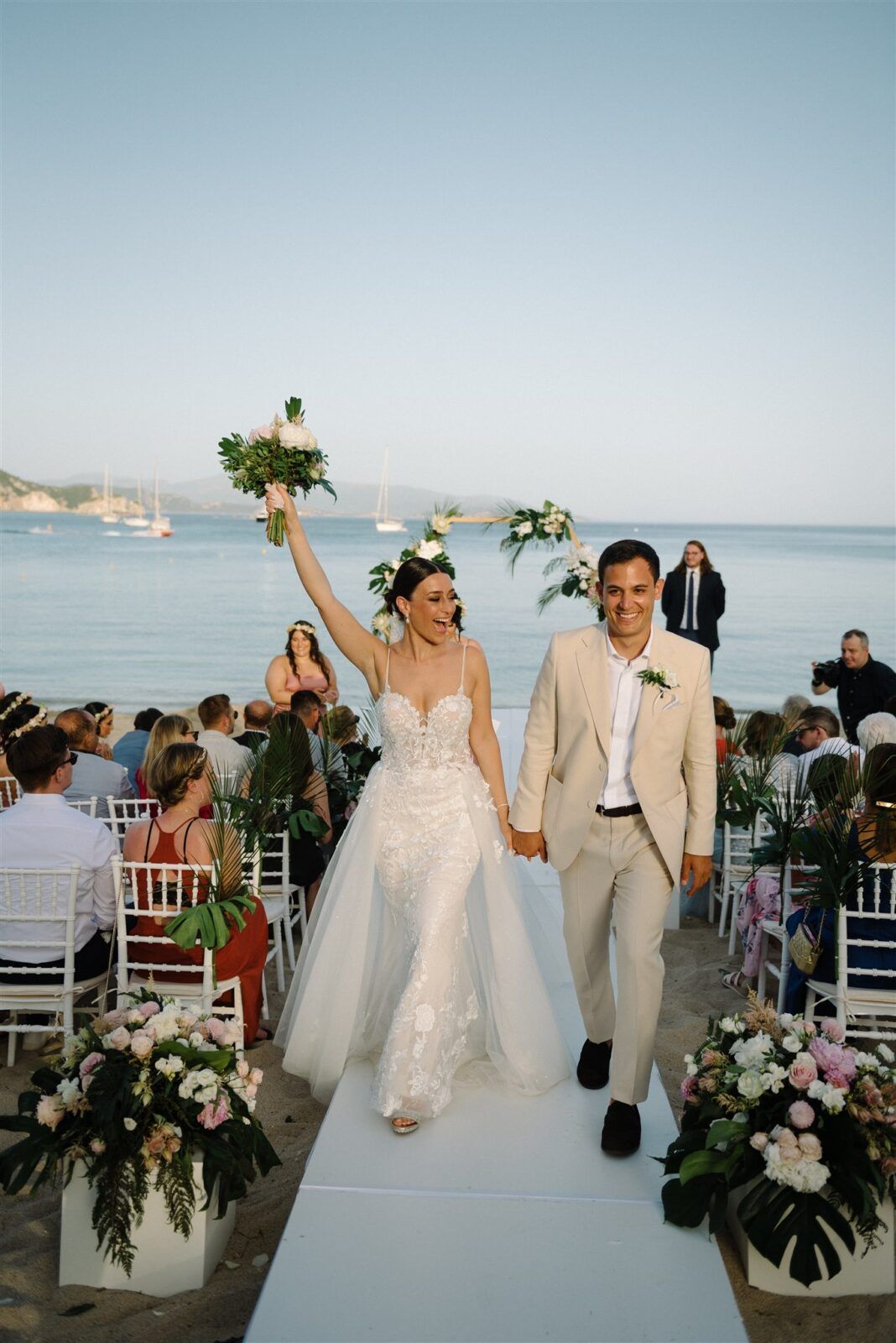 A wedding ceremony on a beachfront with white chairs, floral decorations and boats visible in the ocean background.