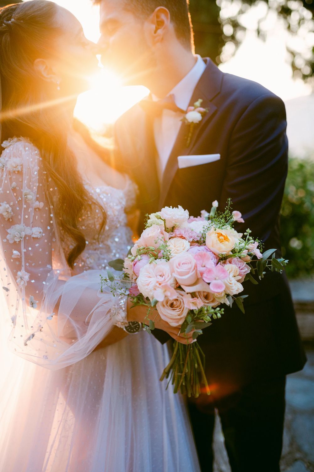 Romantic sunset wedding photos showcase delicate pink and peach rose bouquet with ethereal lighting and flowing white dress details.