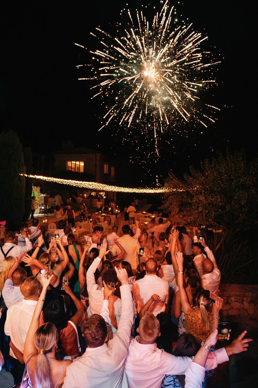 A large crowd dressed in white celebrates under fireworks at an outdoor nighttime event.