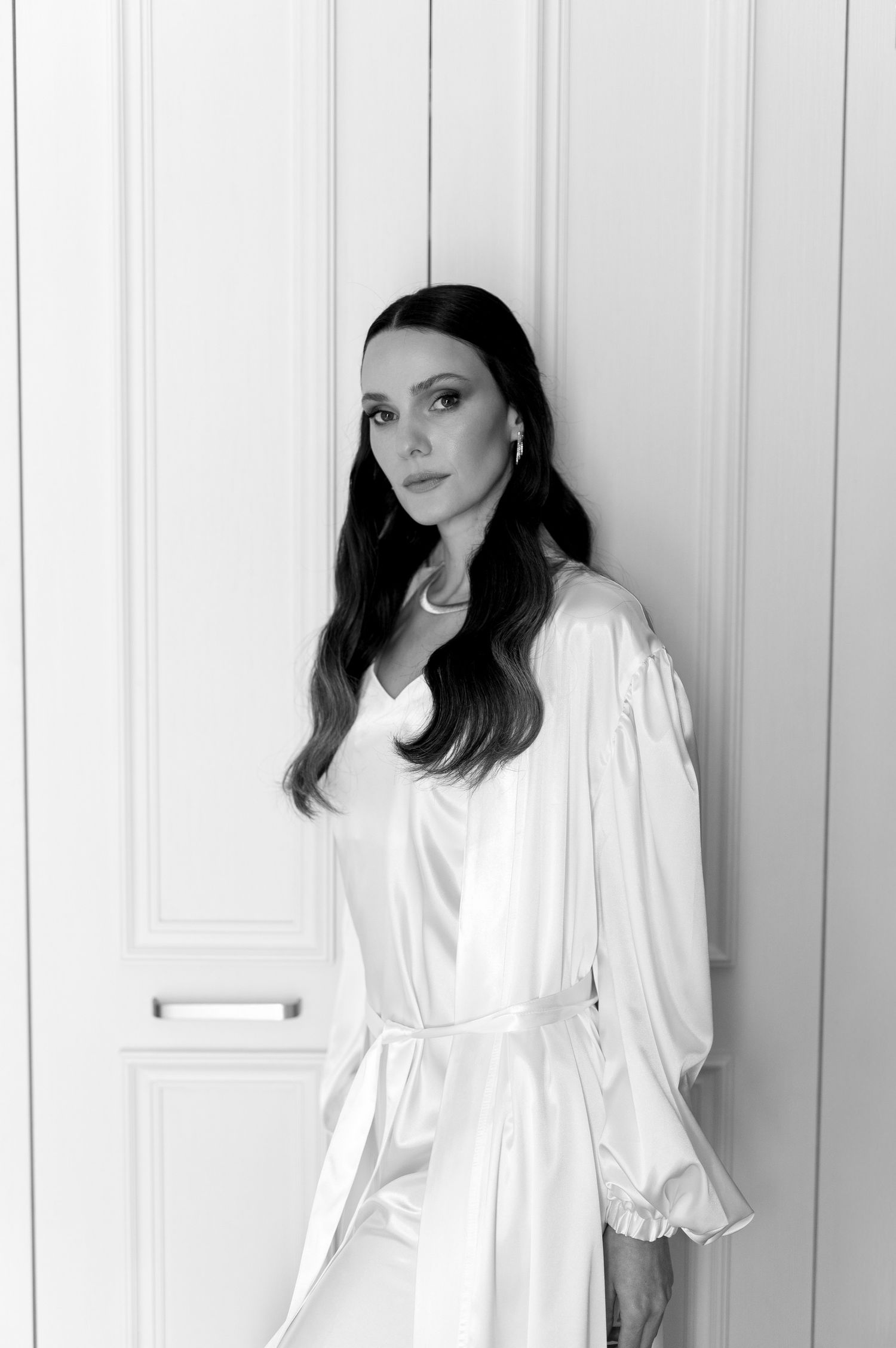 Black and white portrait of someone in a flowing white dress against a light colored wall with paneling.